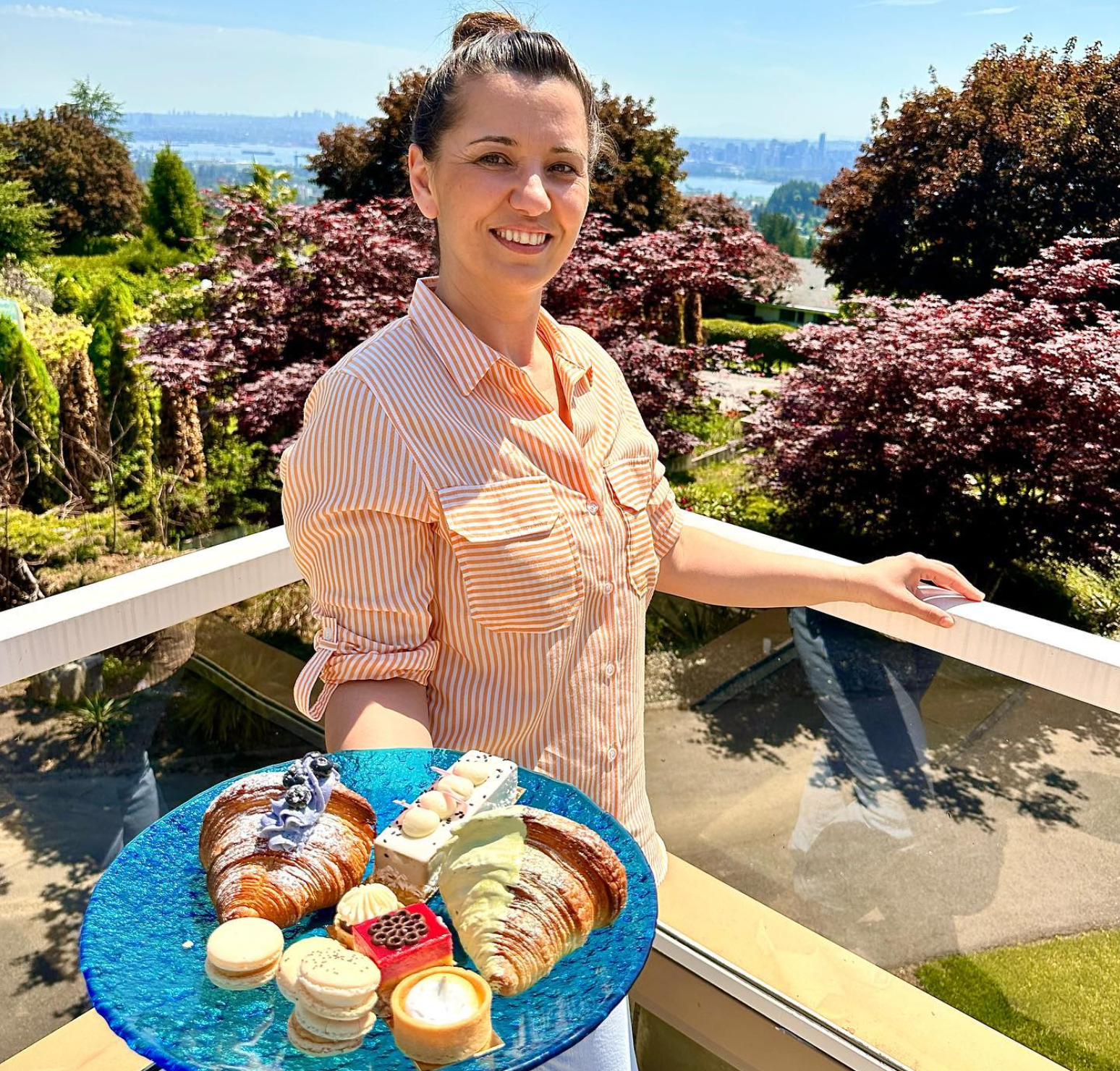 Nena holding a blue plate with assorted desserts, including croissants, macarons, tarts, and other pastries, on a balcony outdoors with trees and a city skyline in the background.