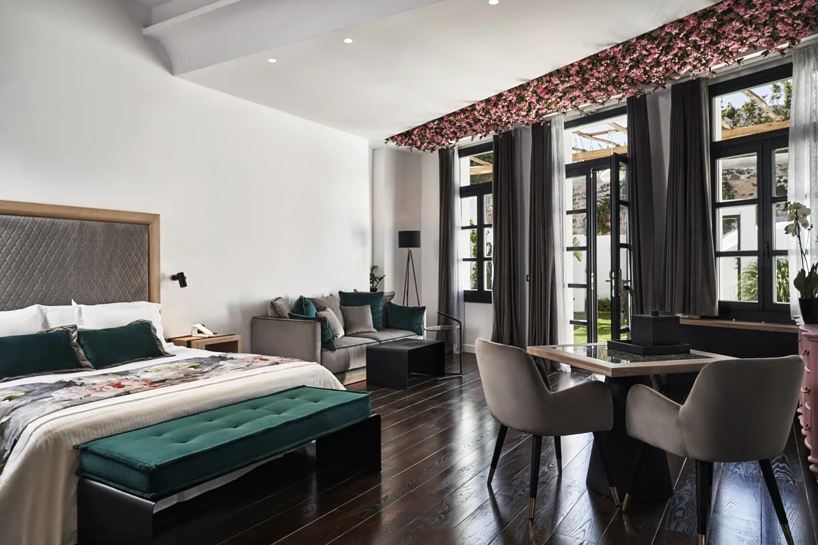 Interior of a modern hotel room featuring a large bed with floral bedding, a gray sofa with teal and gray cushions, a round dining table with beige chairs, and large windows with dark curtains and a view of the outdoors, including a pink flower ceiling decoration.