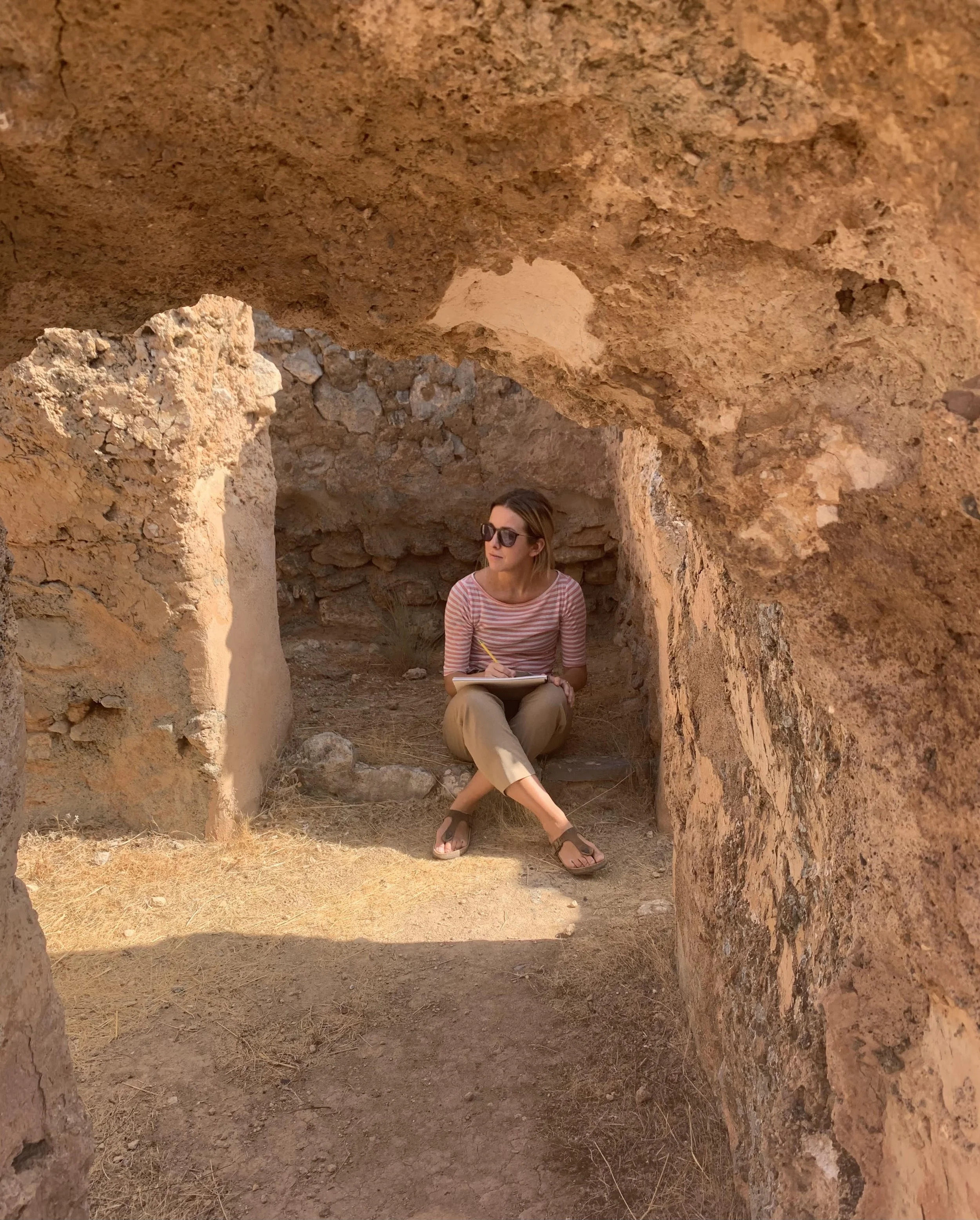 A woman sitting inside a small cave or alcove made of rocks, wearing sunglasses, a striped shirt, and khaki pants, holding a notebook and pencil, with dry ground and rocks around her.