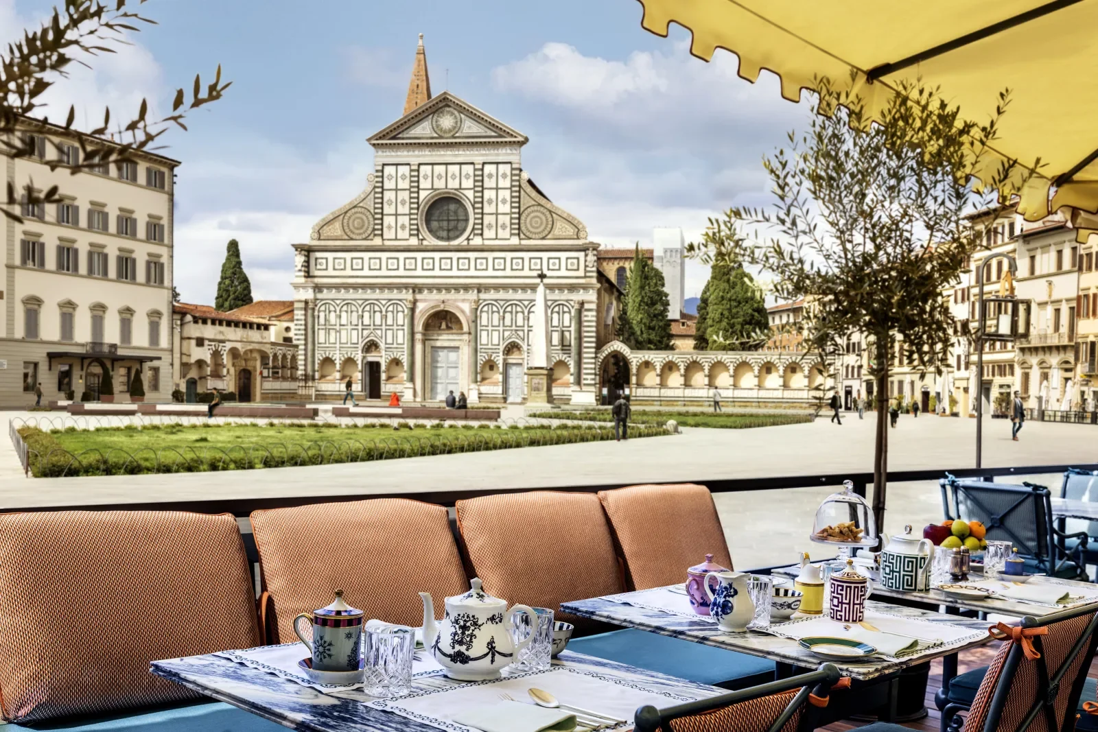 Outdoor café table with patterned teapots, cups, and glasses, overlooking the historic Santa Maria Novella church in Florence, Italy, with a yellow awning and cityscape in the background.