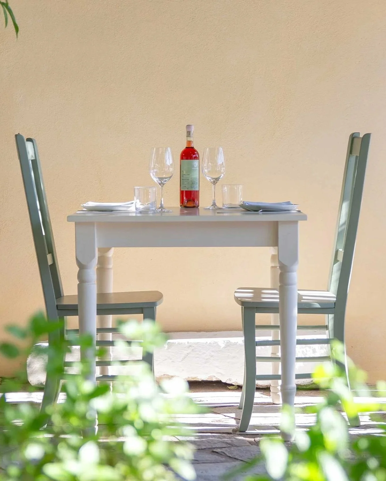 A white outdoor table set for two with wine glasses, a bottle of rosé wine, and plates with napkins, against a yellow wall, with green plants in the foreground.
