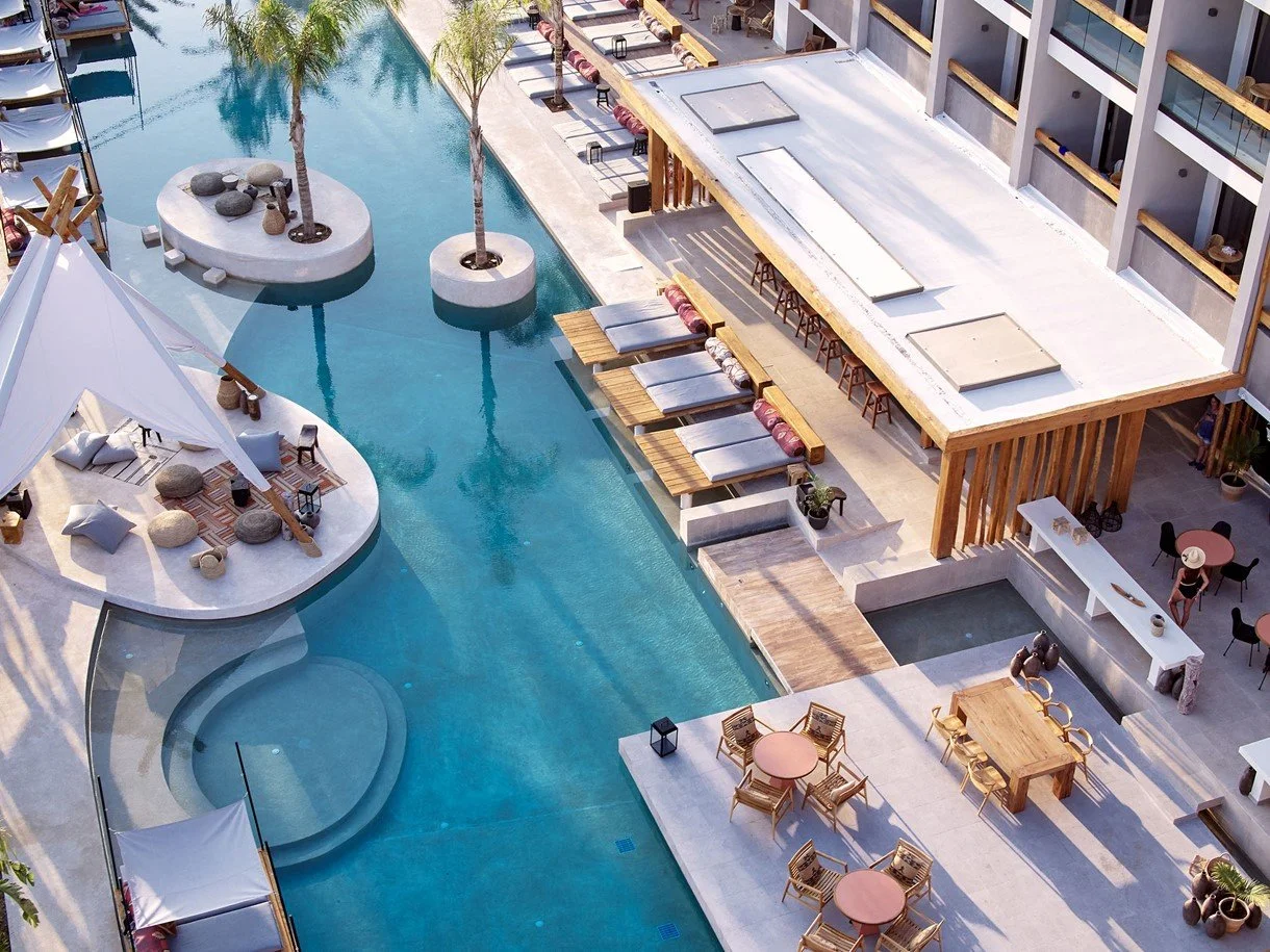 Aerial view of a modern hotel pool area with lounge chairs, palm trees, and surrounding buildings.