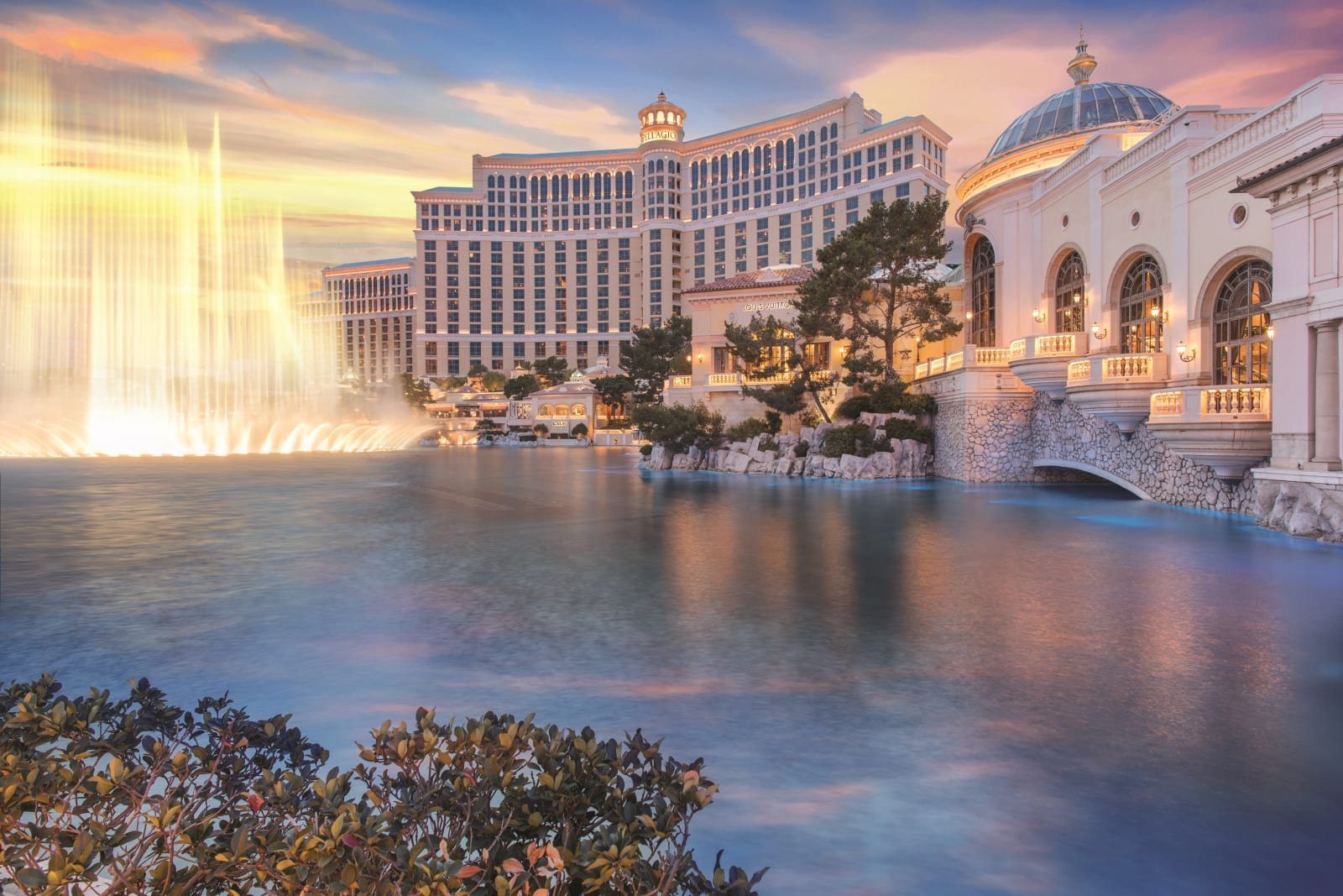 View of the Bellagio Hotel and Casino in Las Vegas at sunset, featuring its fountain show with water spraying, a large lake, and classical architecture with a dome and balcony, under a colorful sky.
