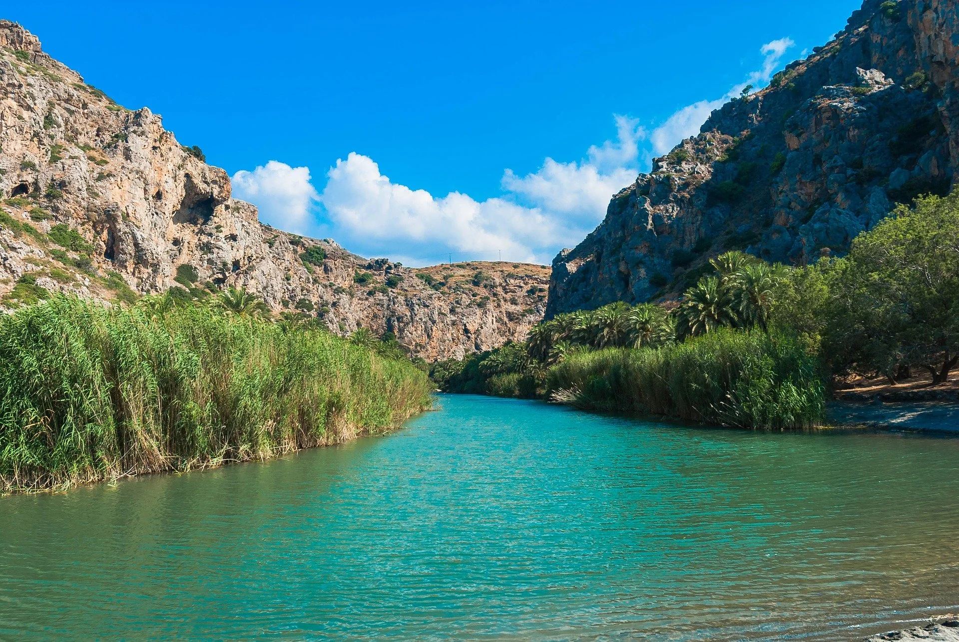 A river flowing between lush green vegetation and towering rocky cliffs under a bright blue sky with white clouds.