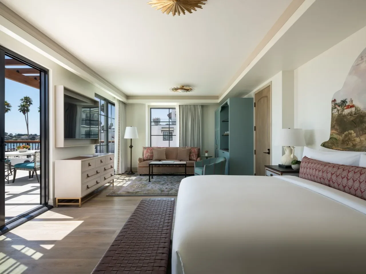 Bright hotel room with a large bed, a sitting area with a sofa and chair, a TV on a white console, and sliding glass doors leading to a balcony with ocean view and palm trees.