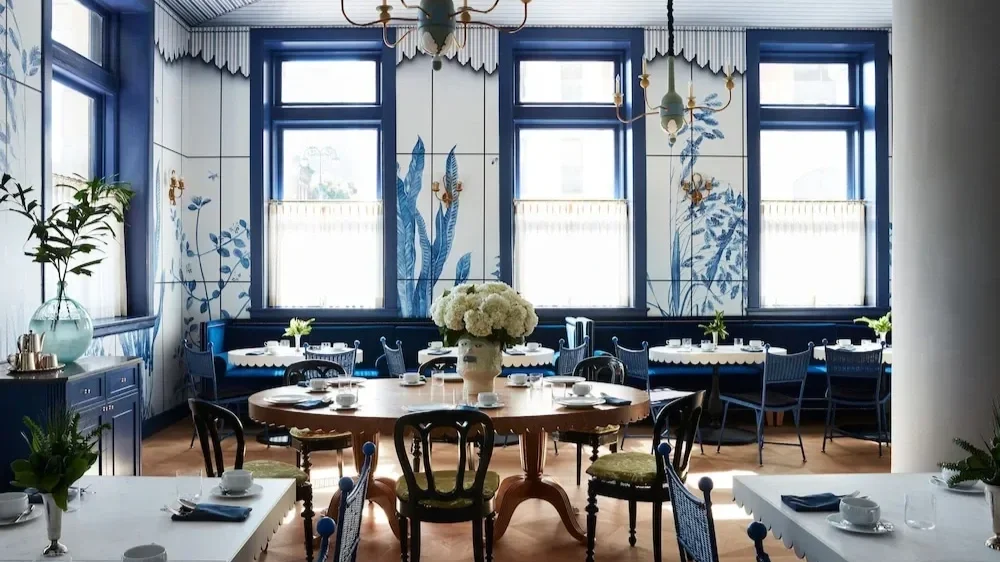 Elegant dining room with blue window frames, floral wall art, and a chandelier, featuring a large central bouquet of white flowers on a round wooden table and other tables set for dining.