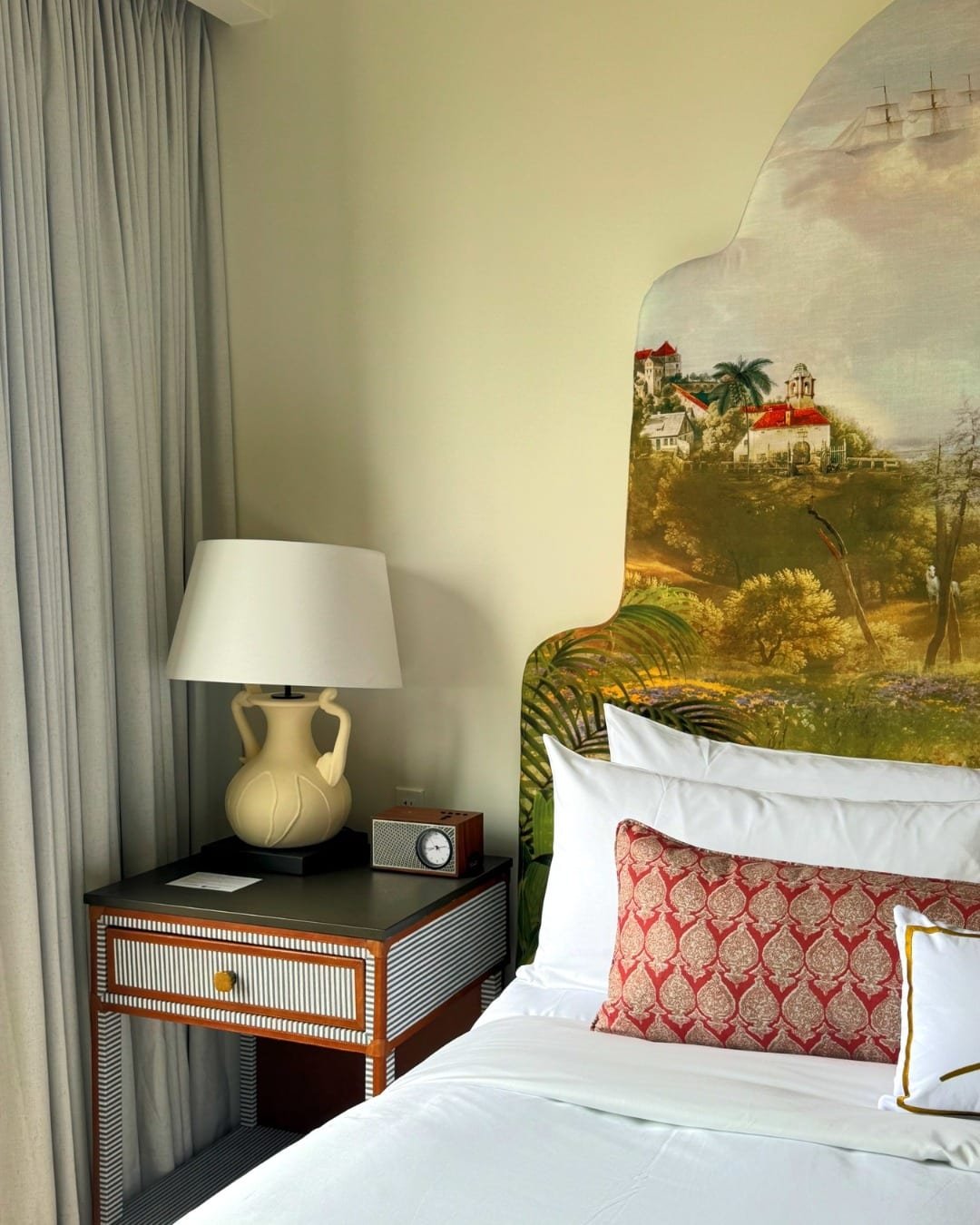 A bedroom corner with a desk, a large lamp, and a bed with decorative pillows, featuring a mural of a scenic landscape on the wall.