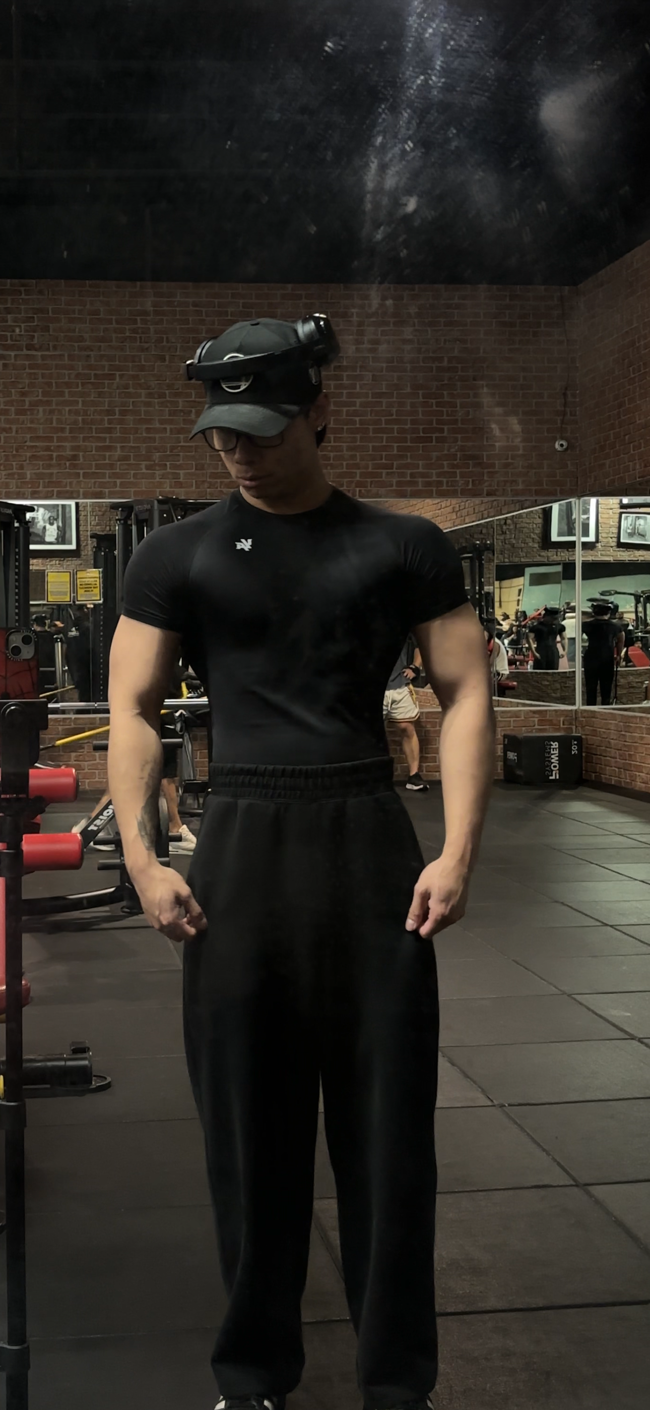 A man in black workout clothes, wearing glasses and a hat with a headlamp, is standing in a gym.