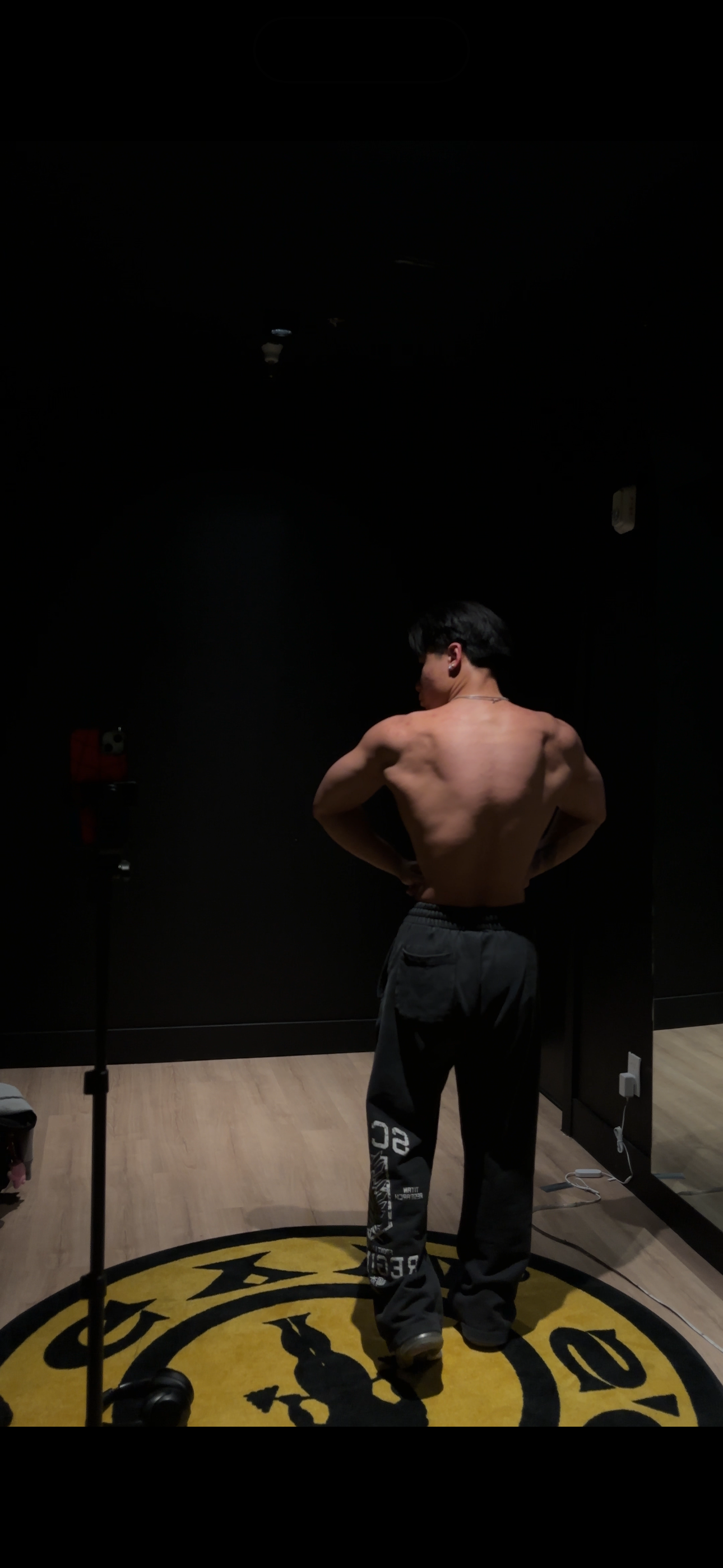 A shirtless man with black hair standing indoors on a circular yellow mat with a black design, facing away from the camera, with dark walls and wooden flooring.