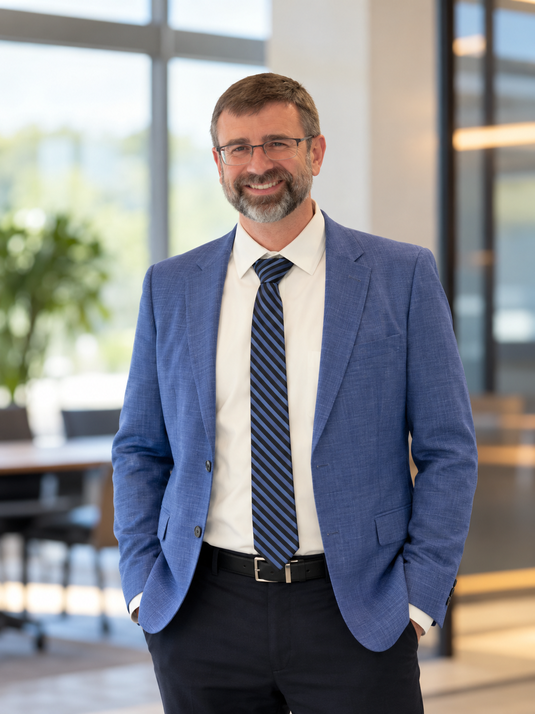 A middle-aged man with glasses, gray beard, and short brown hair, wearing a blue suit, white shirt, and striped tie, smiling and standing with his hands in his pockets in an office setting.
