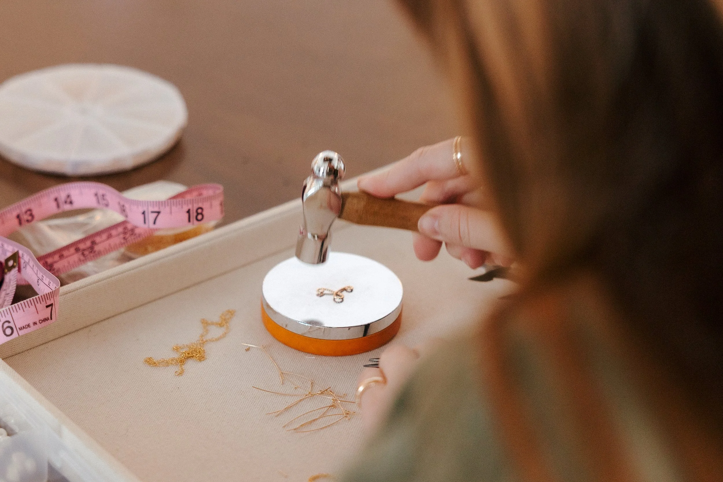 Person placing a small jewelry piece on a white circular display stand with a wooden surface, a pink measuring tape, and gold jewelry chains nearby.