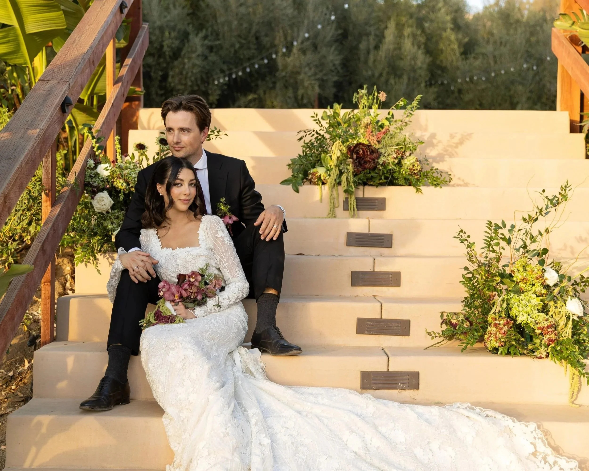 A couple dressed in wedding attire sitting on outdoor stairs with greenery and floral arrangements around them, during sunset.
