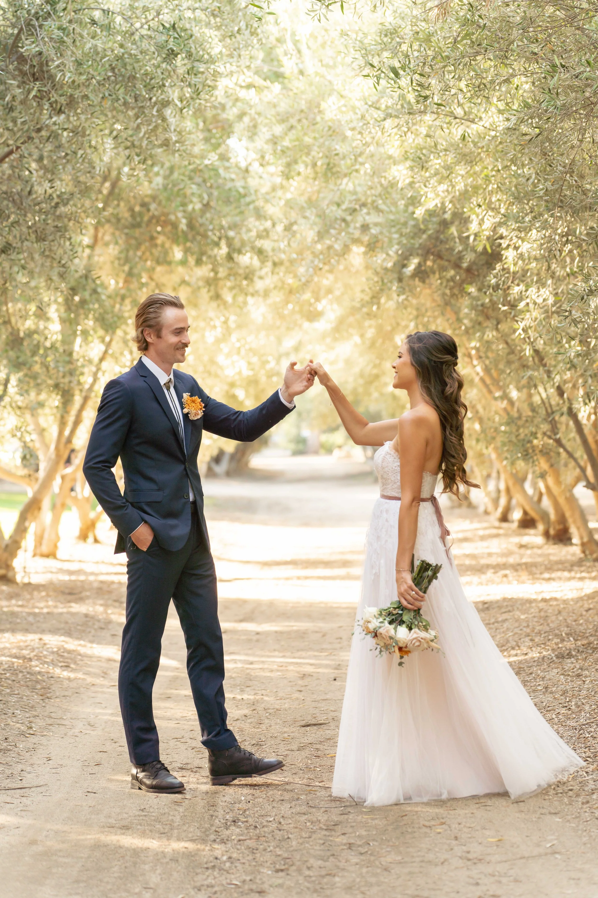 A bride and groom are outdoors, exchanging a fist bump. The groom is in a dark suit with a flower boutonnière, and the bride is in a strapless white wedding dress holding a bouquet of flowers. They are surrounded by a pathway lined with trees.