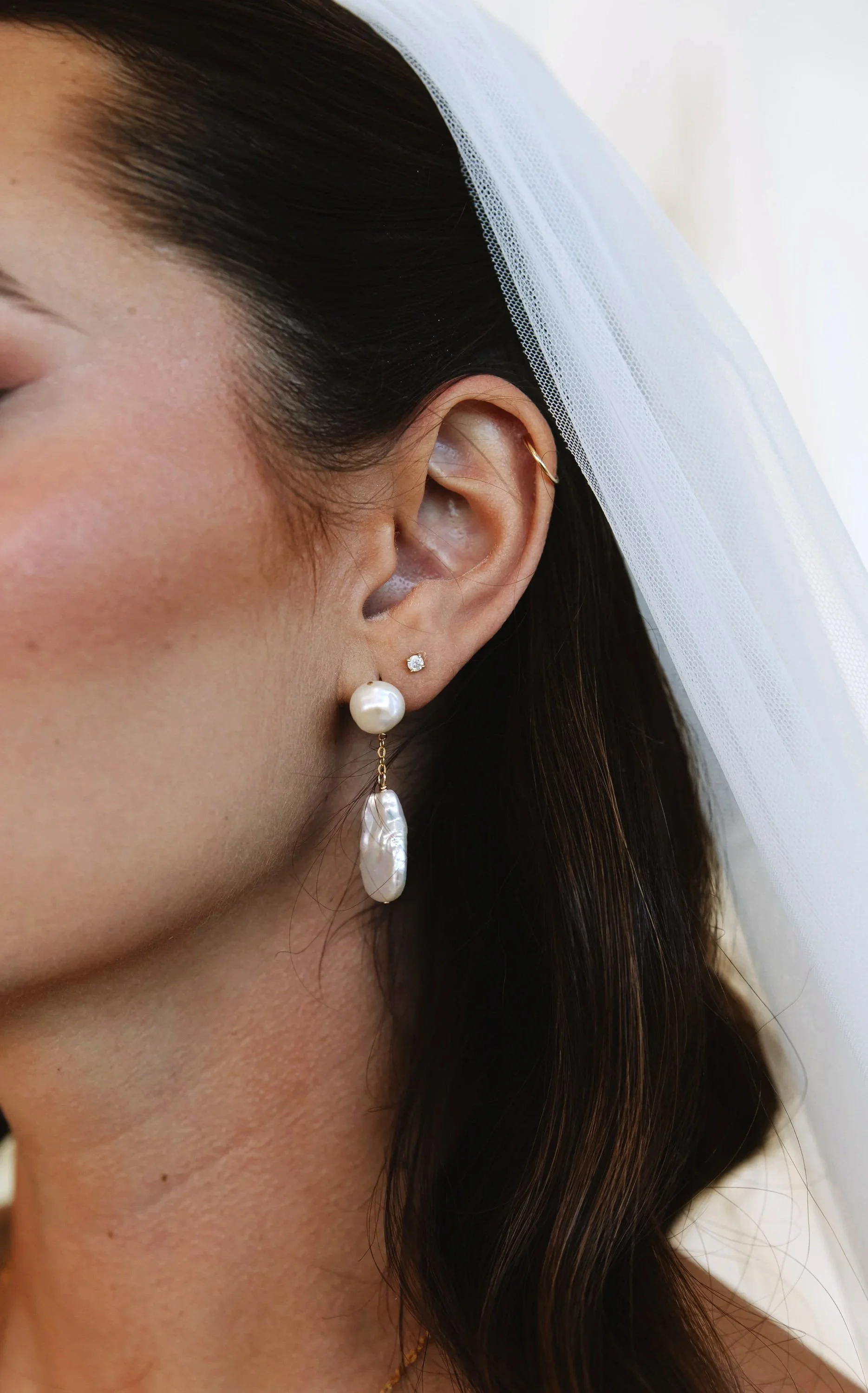 Close-up of a woman's ear with three earrings: a small diamond stud, a small hoop, and a large pearl drop earring, with dark brown hair and white background.