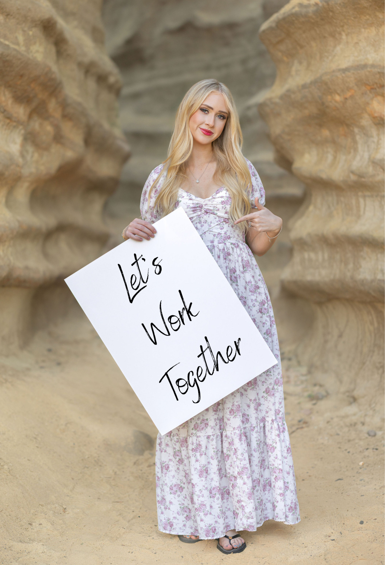 Portrait of a young woman in a floral dress holding a sign that reads "Let's work together" at a rocky outdoor location.