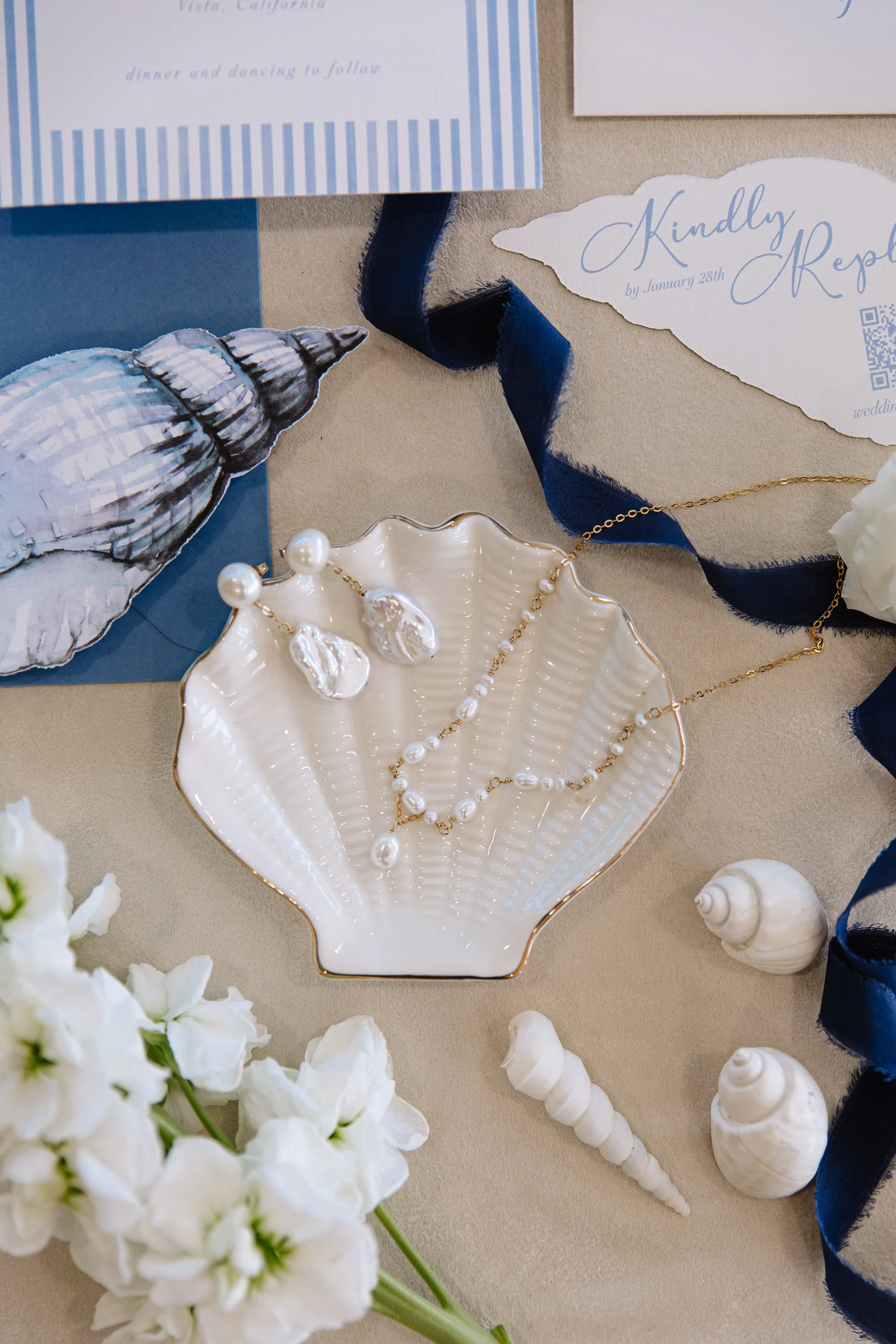 Decorative seashell-shaped dish with pearl and gold jewelry, surrounded by white flowers, seashells, and invitations with calligraphy on a beige surface.