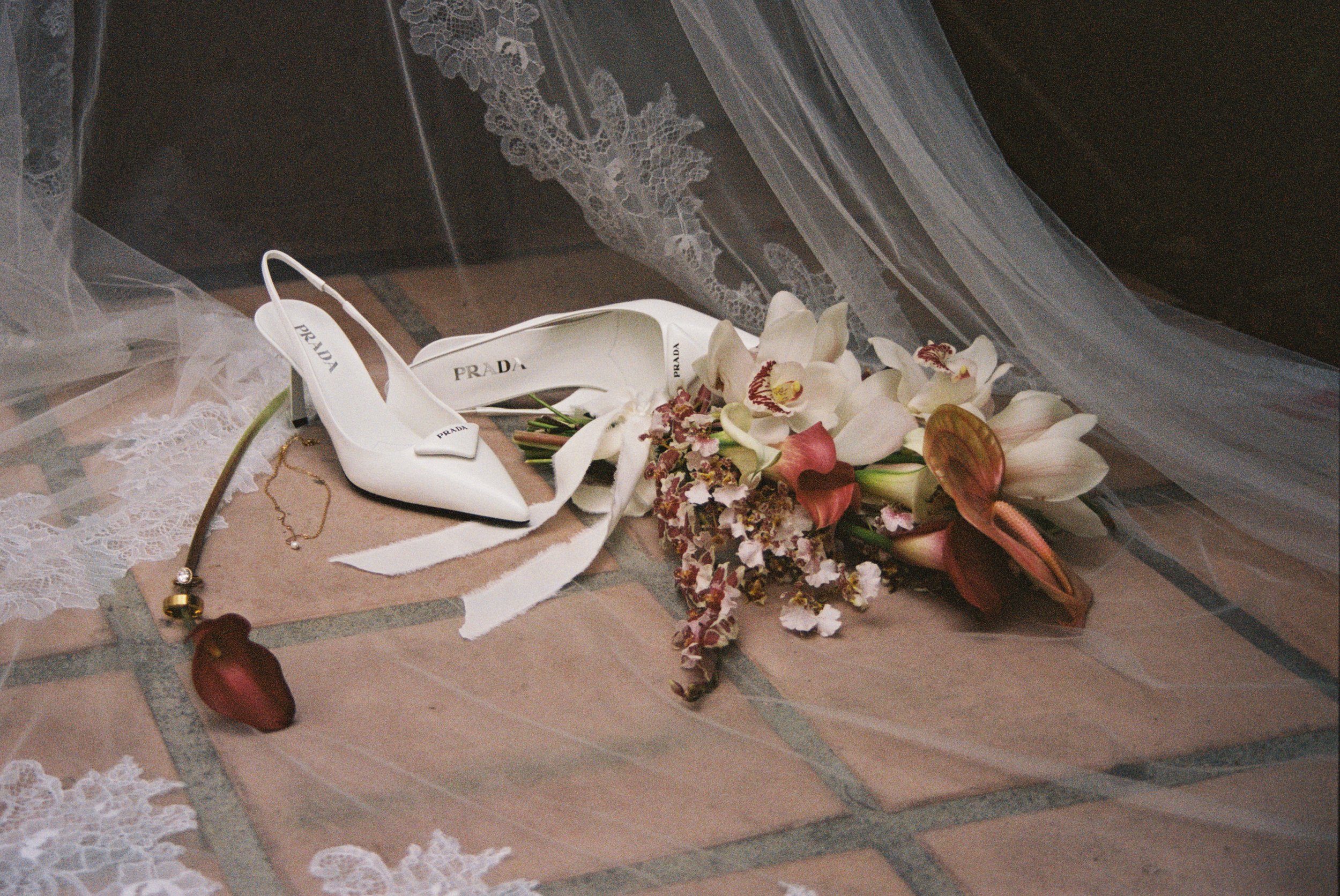 Prada white high-heeled shoes surrounded by flowers and jewelry on a tiled floor, with lace and tulle fabric nearby.