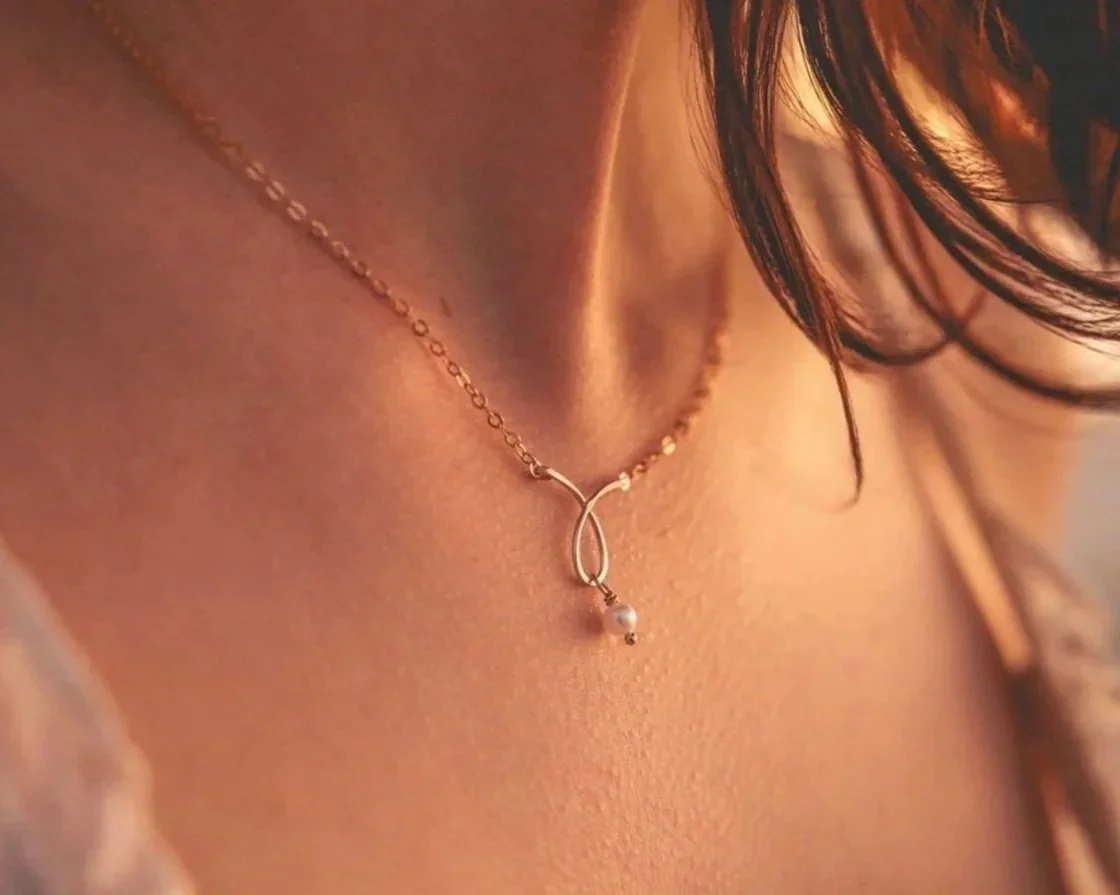 Close-up of a woman's neck wearing a gold necklace with a teardrop-shaped pendant and a small pearl.
