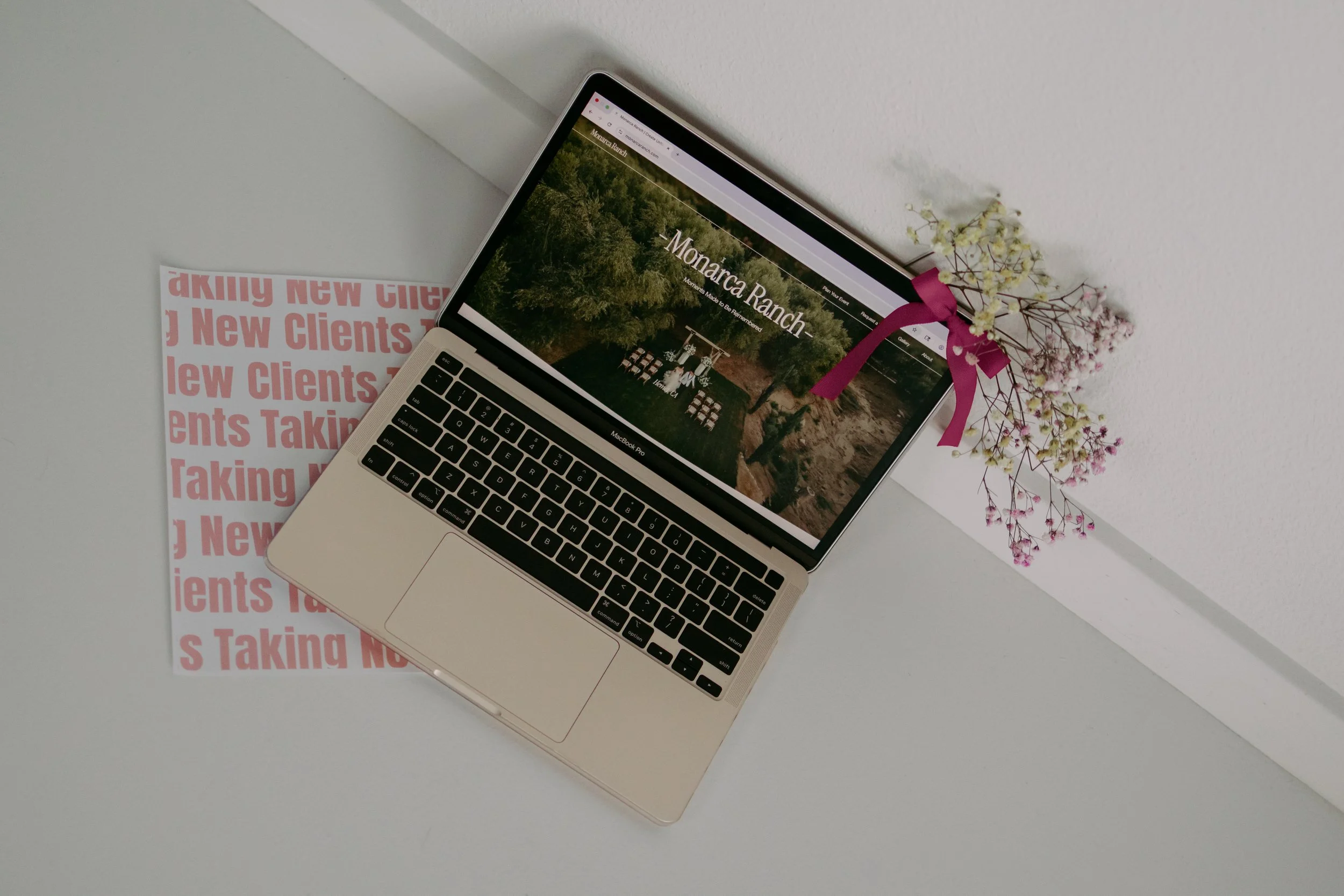 A silver laptop displaying the website 'Monarcha Ranch' with a background of green trees. A small bouquet of artificial flowers with pink and white blooms tied with a pink ribbon is placed behind the laptop. To the left, there is a partially visible pink and white printed paper with repeated text 'New Clients Taking' on a white surface.