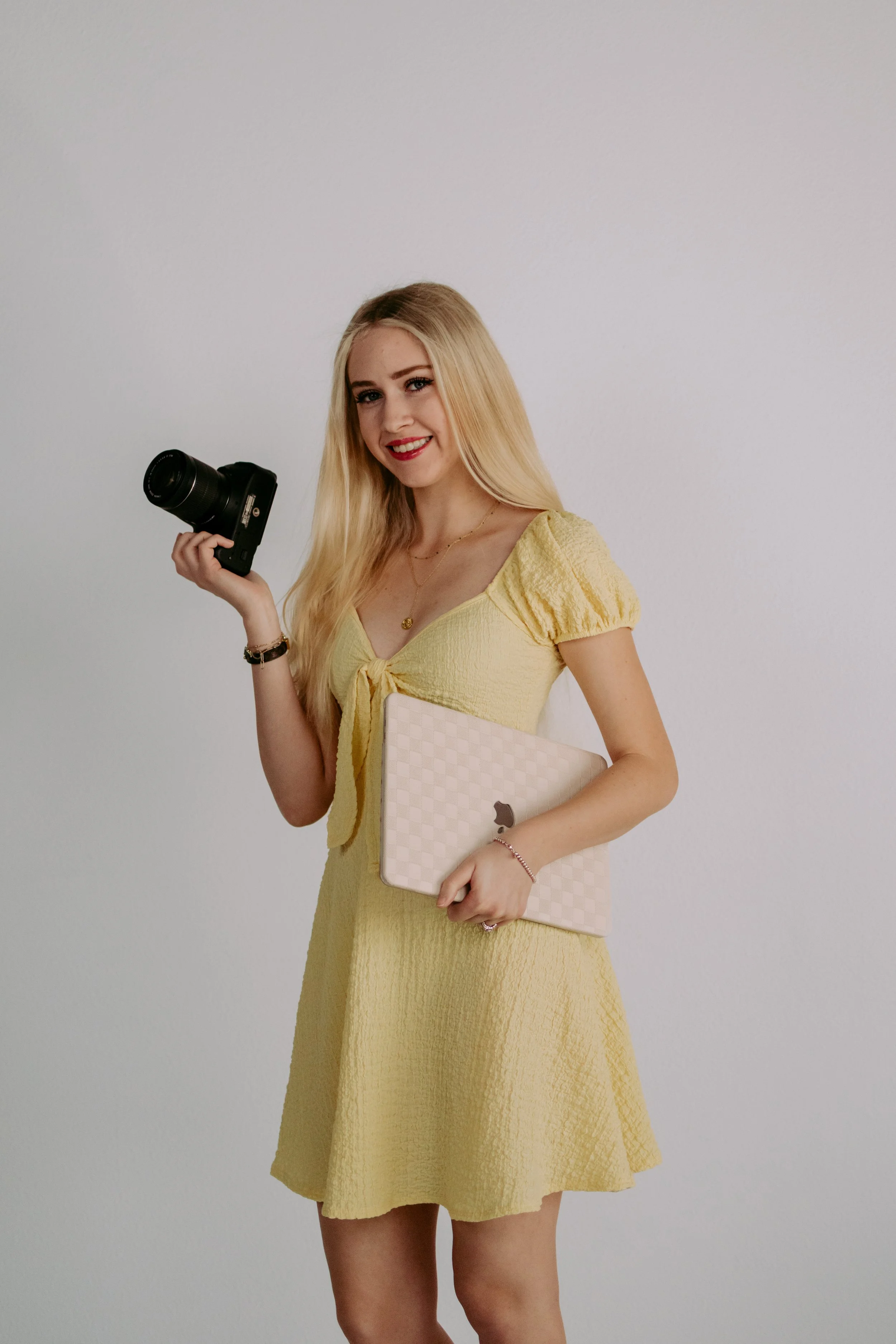 Young woman with blonde hair in a yellow dress holding a camera in her right hand and a pink checked laptop in her left hand against plain white wall.