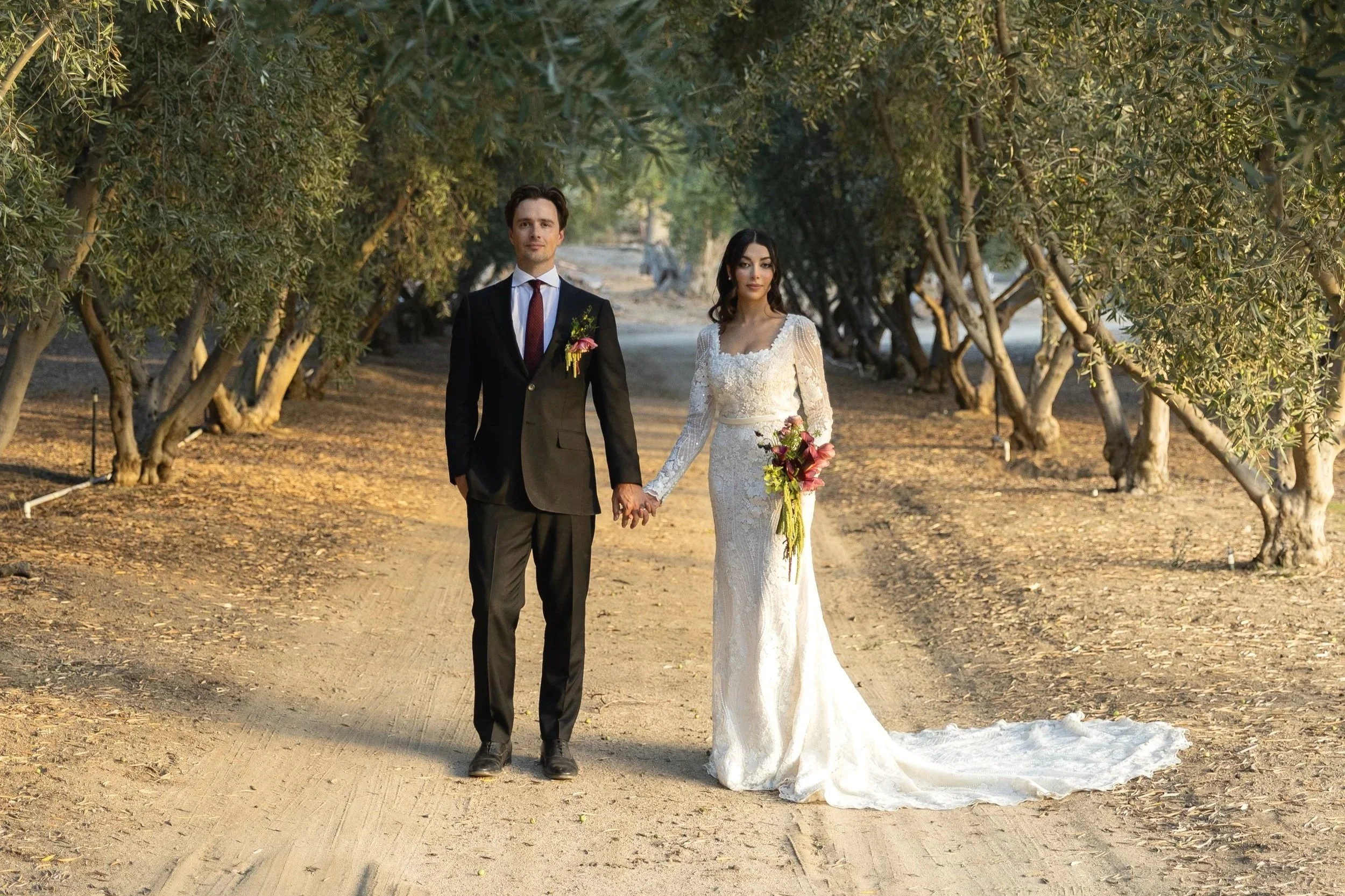 A bride and groom walk hand-in-hand through a tree-lined outdoor setting, dressed in wedding attire, with the bride holding a bouquet of flowers.