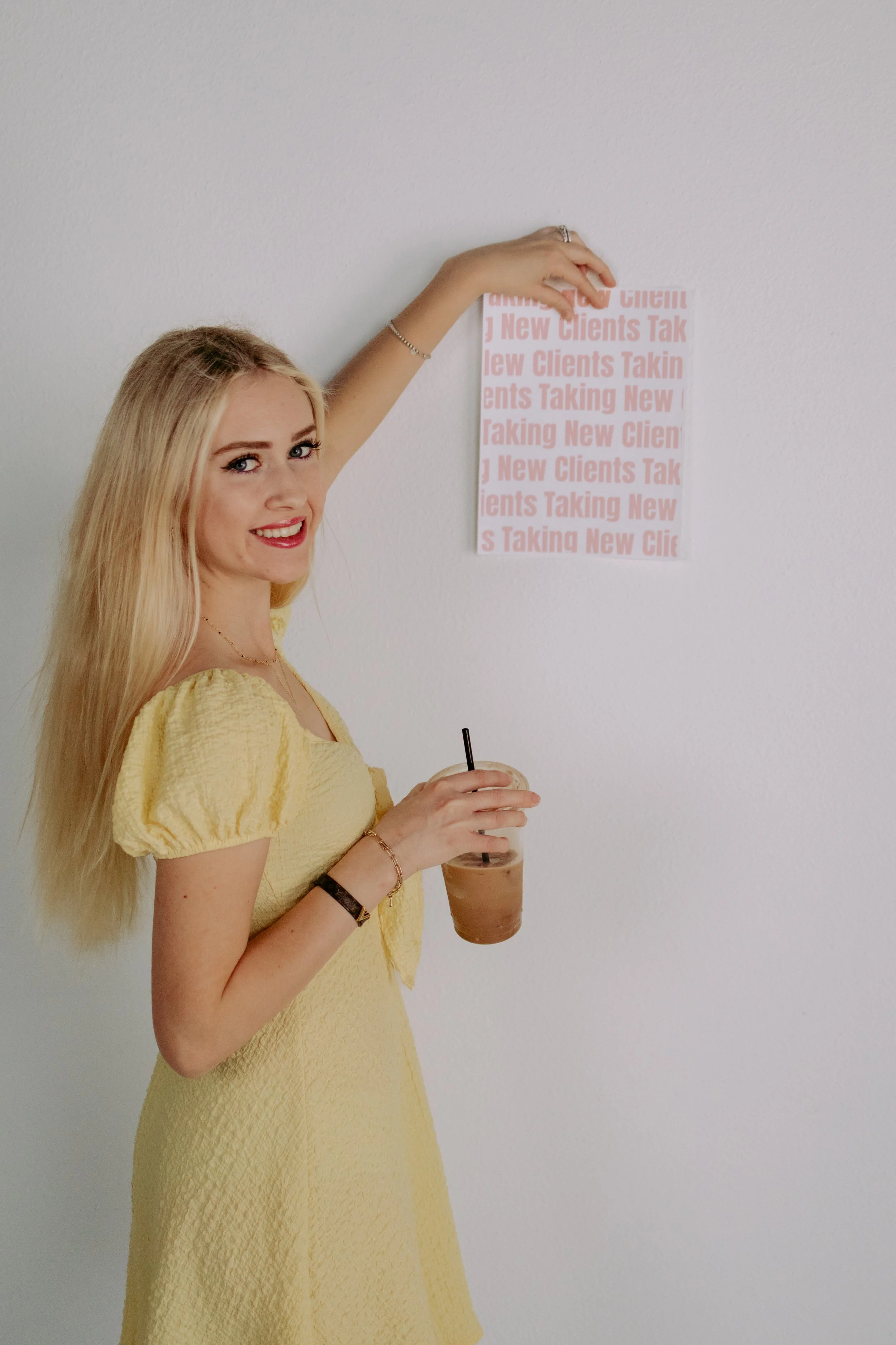 A woman with long blonde hair in a yellow dress holds an iced coffee and stands in front of a white wall with a pink poster that repeats 'New Clients Taking' text.