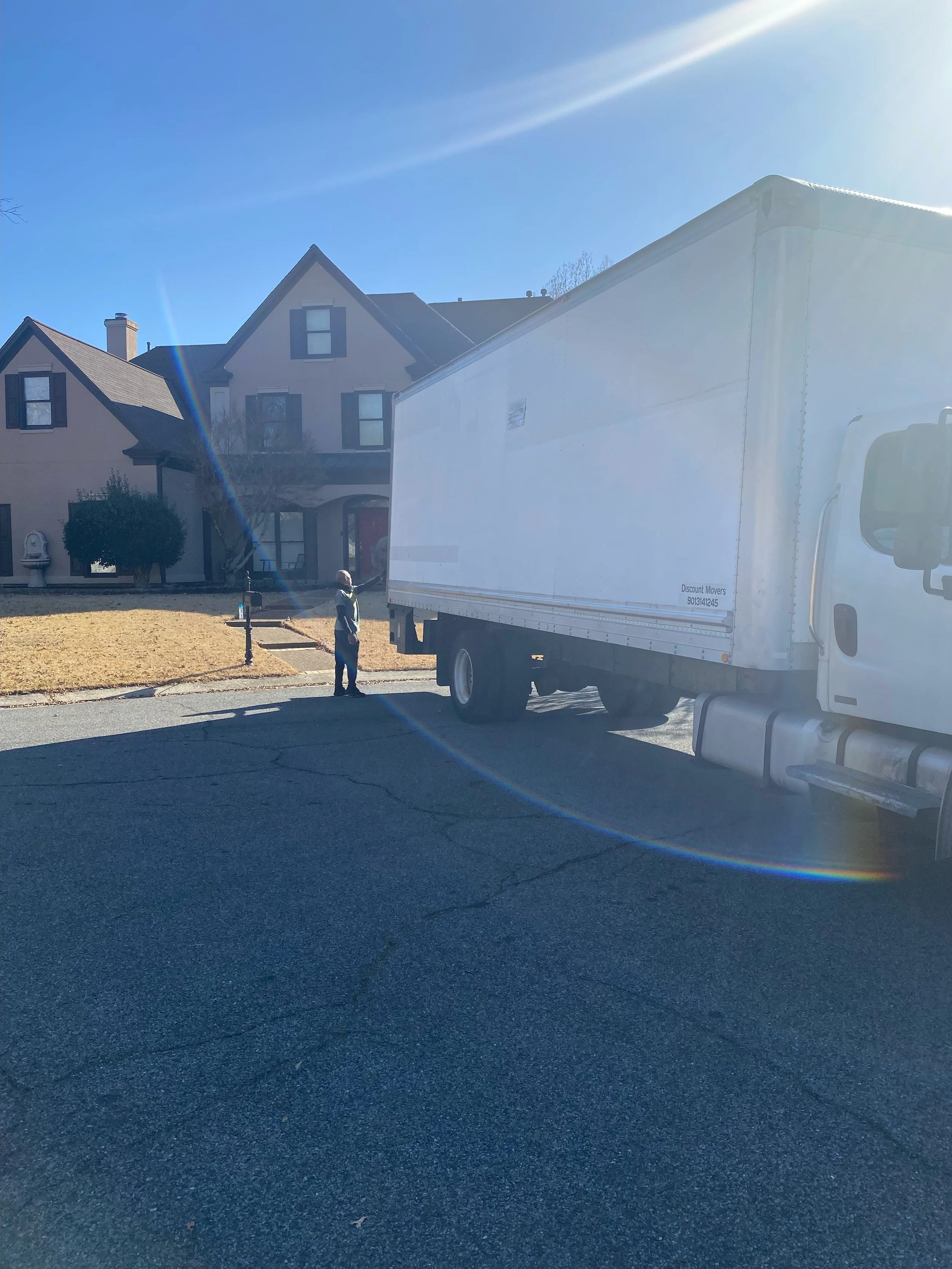 A white moving truck parked in a neighborhood street under a clear blue sky with sunlight. Two houses with front yards are visible, and a person is standing near the truck.