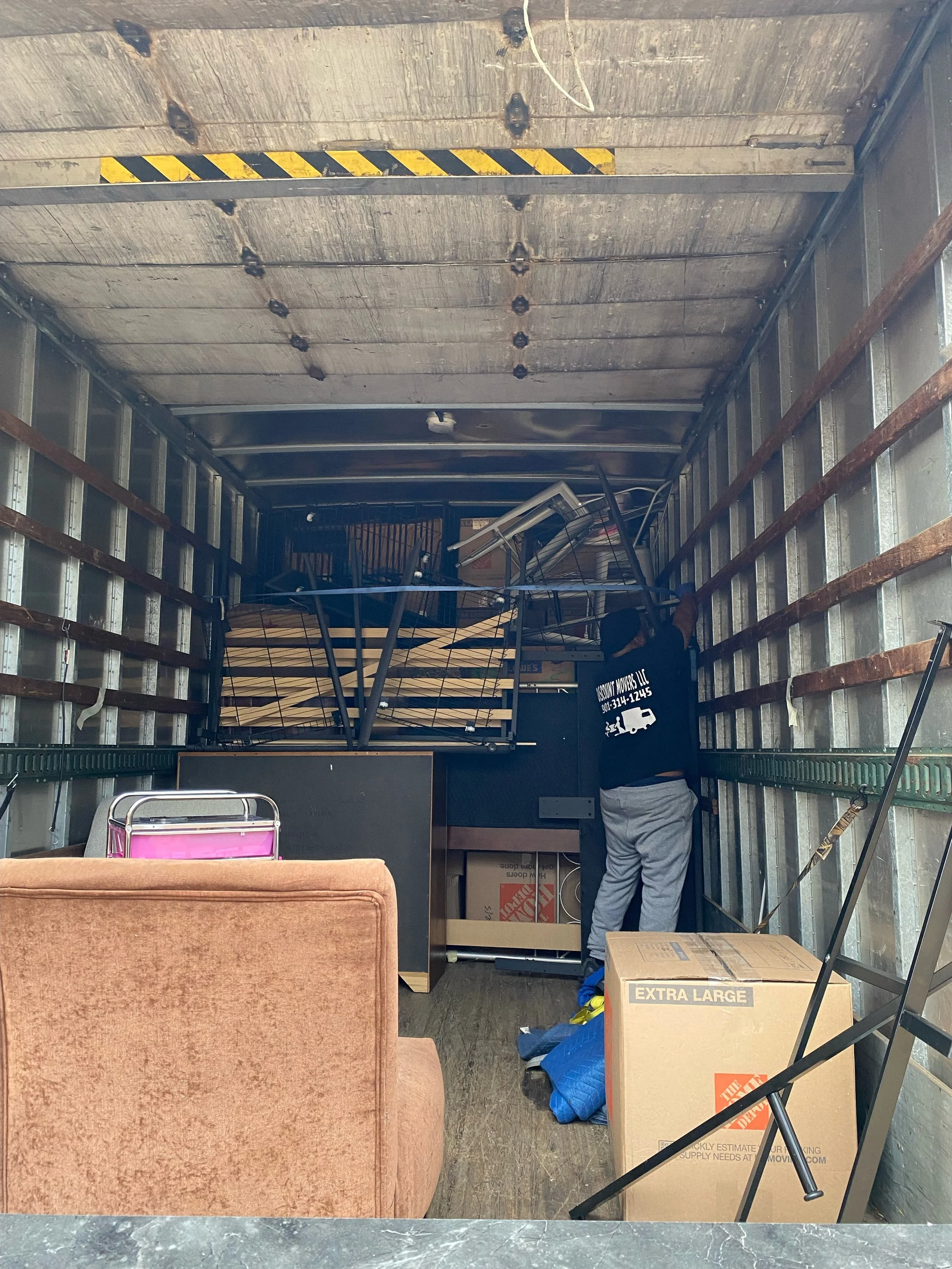Interior of a moving truck filled with furniture and boxes, including a brown upholstered chair, a pink suitcase, and a cardboard box labeled 'Extra Large'; a person in gray pants and a black shirt is organizing items near the back of the truck.