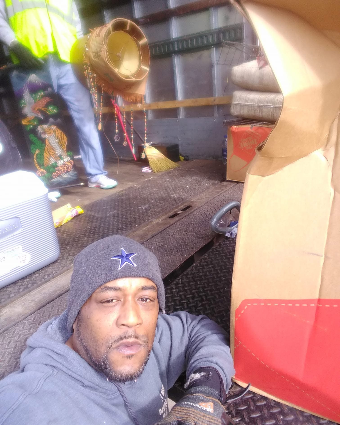 A man wearing a gray beanie with a star logo taking a selfie inside a truck or trailer. The background shows various items including a yellow hat, a broom, and some cushions or mats, with a worker wearing a high-visibility vest partially visible.