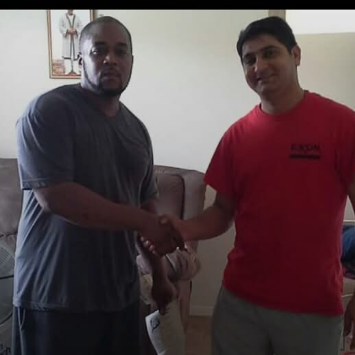 Two men shaking hands indoors, one wearing a black T-shirt and the other in a red T-shirt.