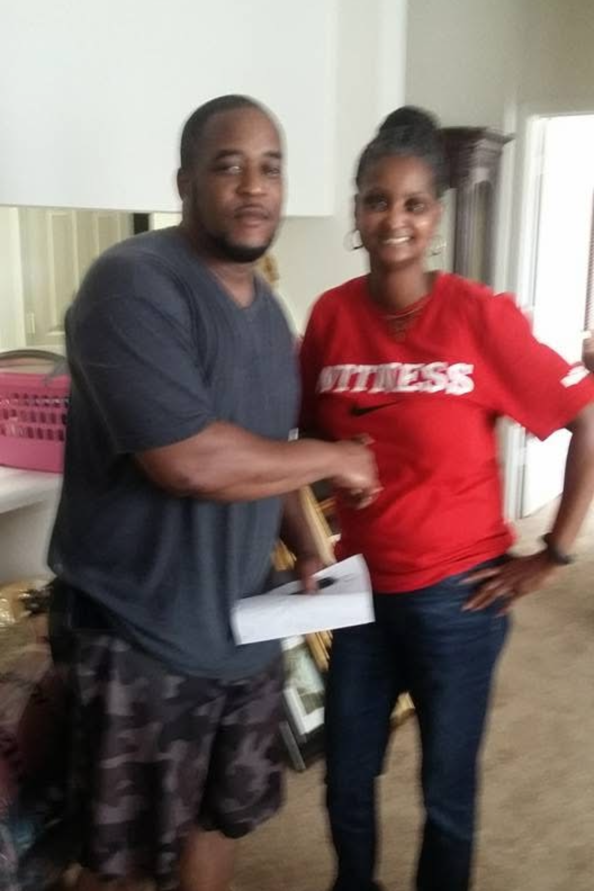 Two people, a man and a woman, standing indoors. The man is wearing a dark t-shirt and patterned shorts, and the woman is wearing a red Nike t-shirt with the word 'FITNESS' on it. They are smiling and appear to be in a casual setting.