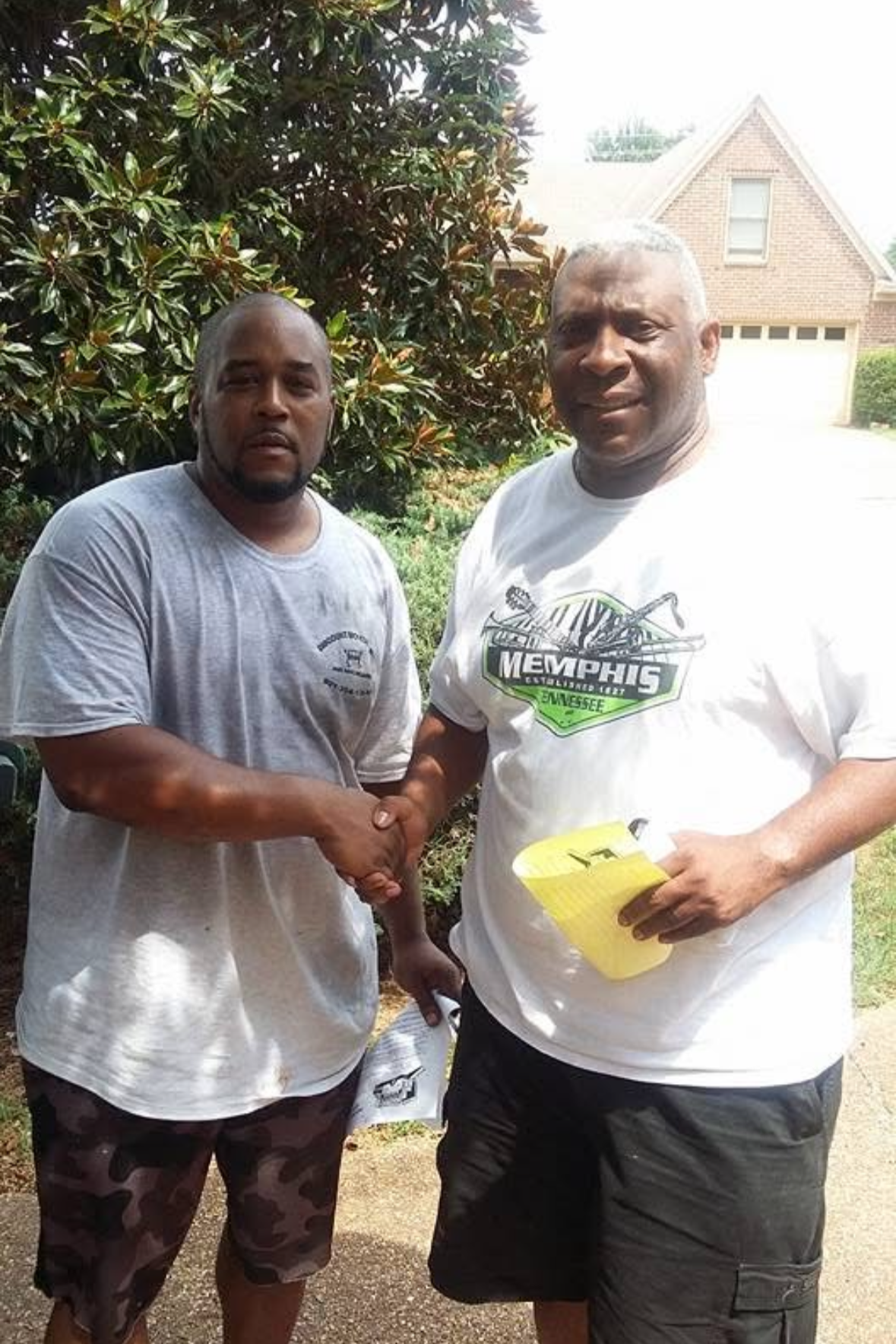 Two men shaking hands outdoors in front of a bush and a house, one holding a yellow container.