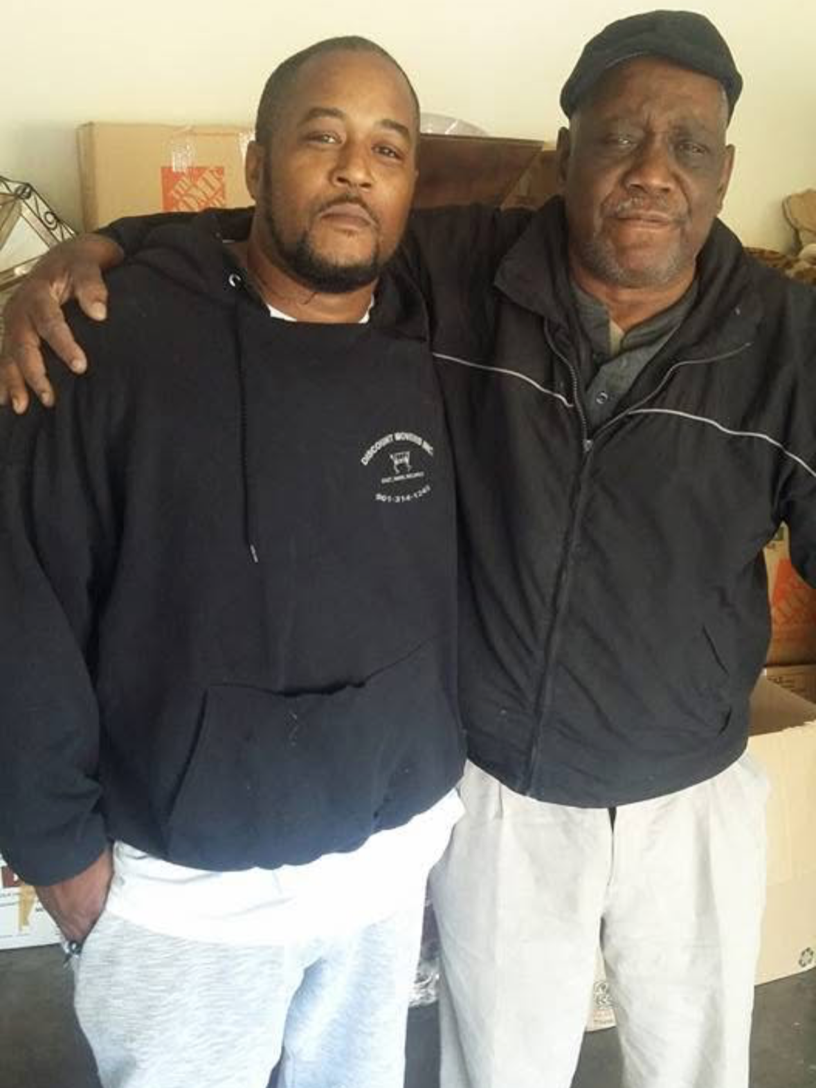 Two men standing side by side indoors, with their arms around each other's shoulders, smiling slightly at the camera. One man is wearing a black hoodie and white sweatpants, and the other is wearing a black jacket with white pants and a hat.