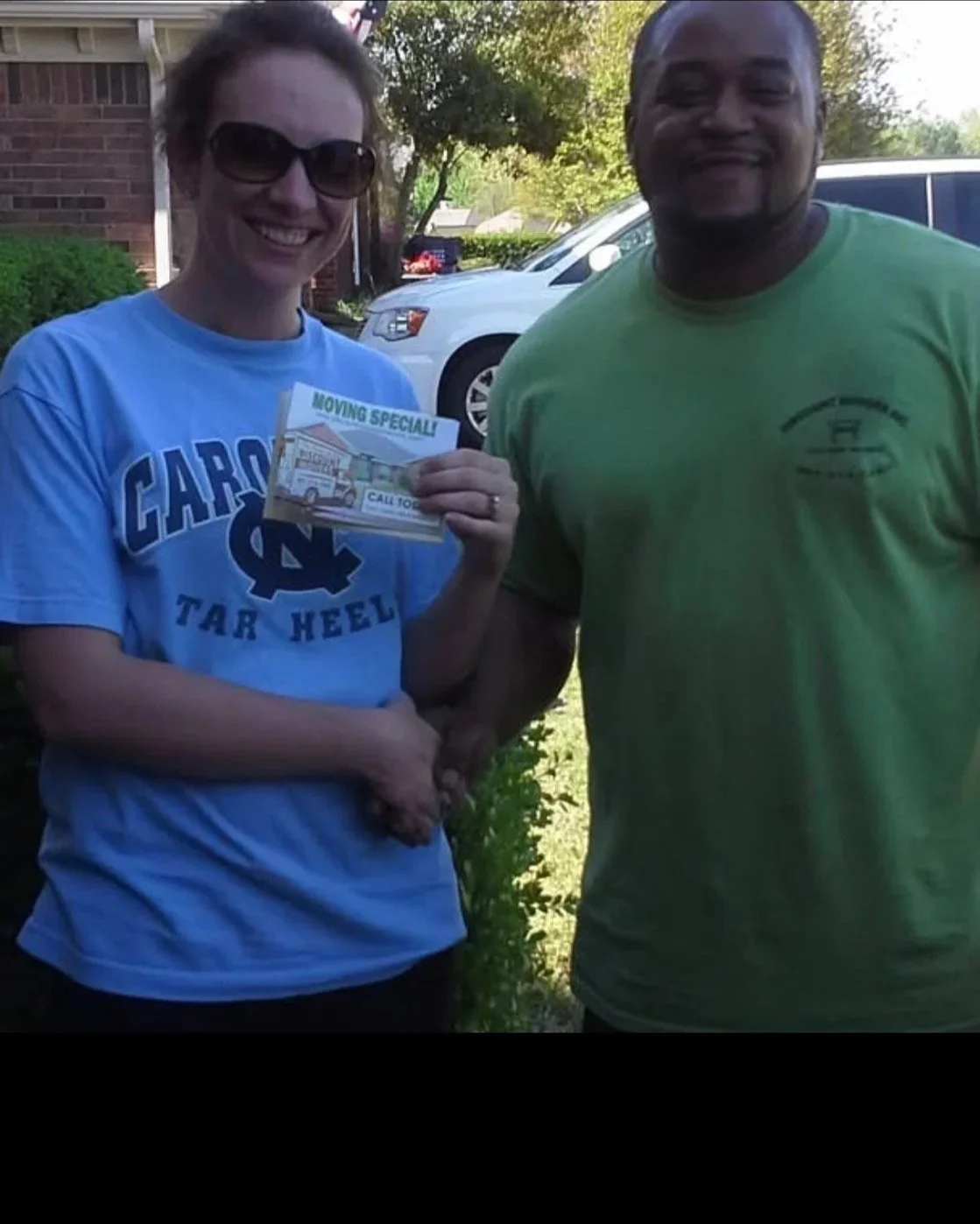 A woman wearing dark sunglasses and a blue 'Carolina Tar Heels' t-shirt holding a flyer for moving specials, shaking hands with a man in a green t-shirt, standing outdoors with cars and trees in the background.