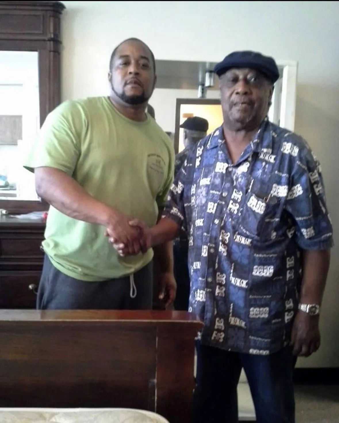 Two men shaking hands in an indoor setting, with one man wearing a green T-shirt and the other wearing a patterned button-up shirt and cap.