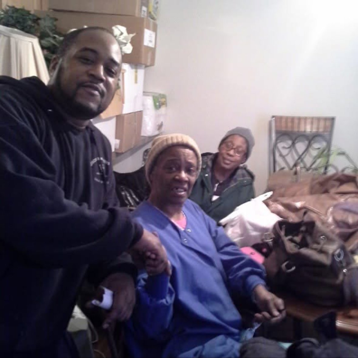 Three people, two women and one man, sit and stand around a table with bags and supplies in a room, with boxes and a wall in the background.