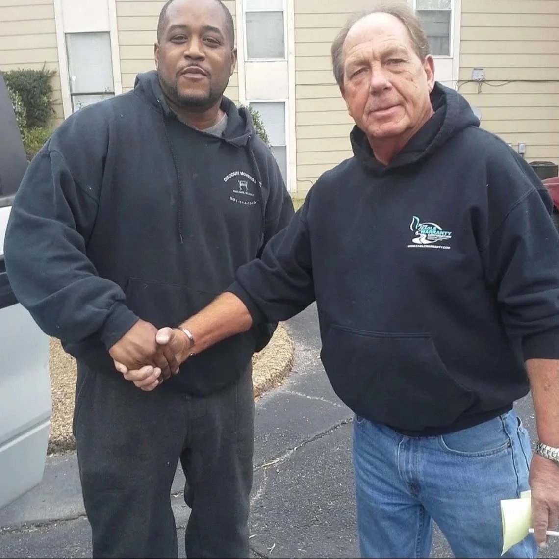 Two men shaking hands outdoors in front of a beige building, both wearing black hoodies with logos.