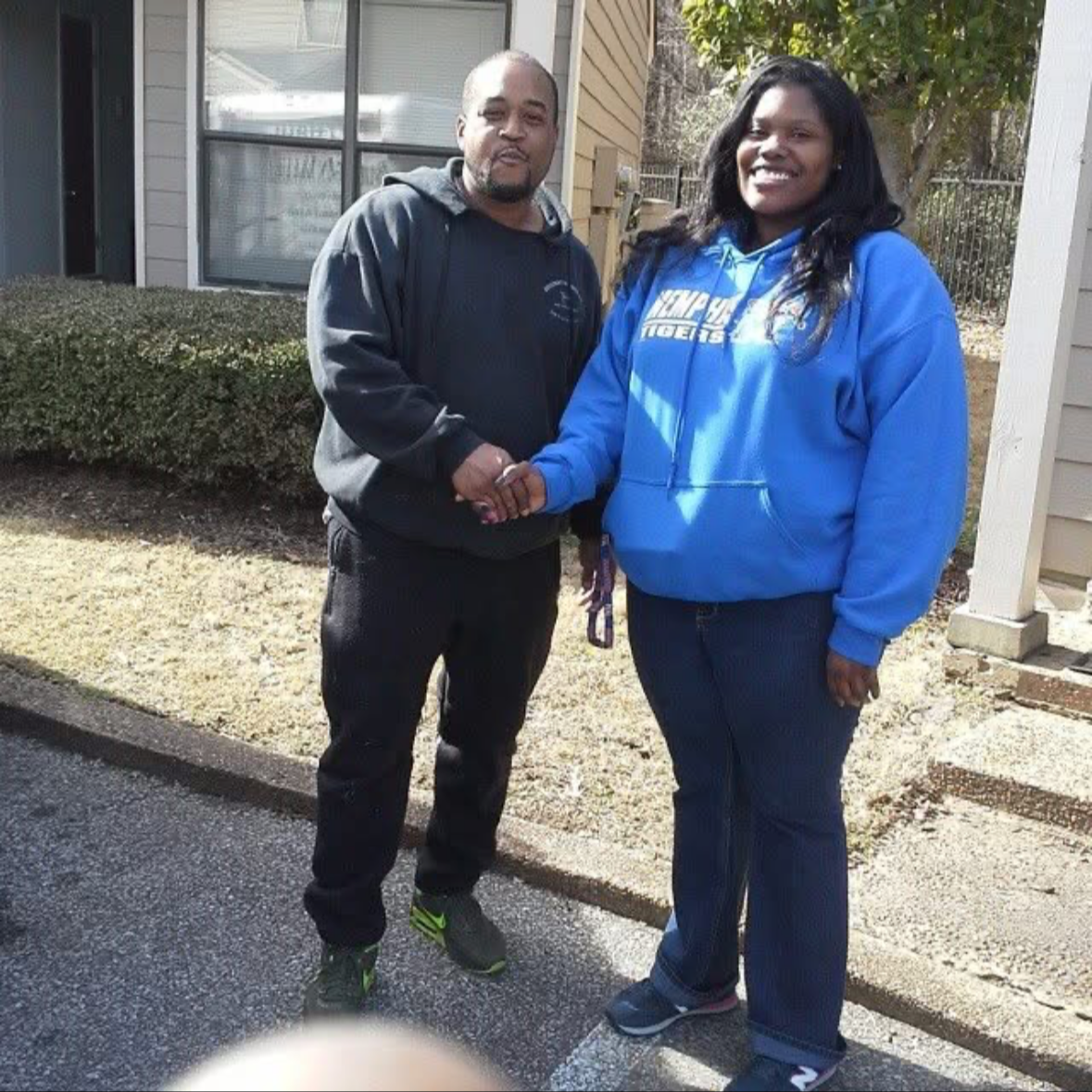 Two people shaking hands outside a building, smiling, with bushes and a fence in the background.