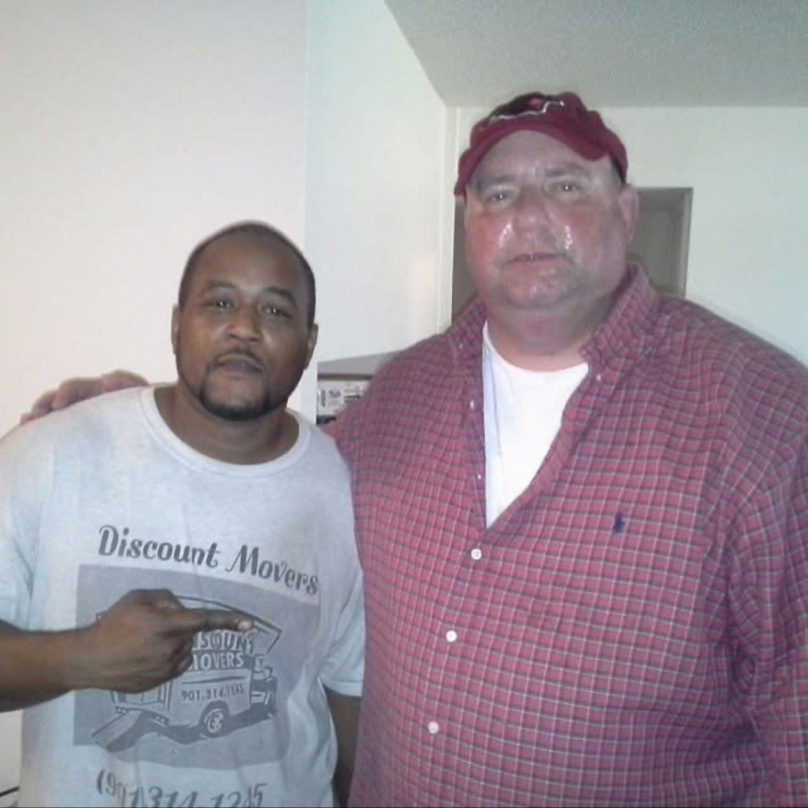 Two men standing close together indoors, one wearing a white t-shirt with a logo and text, the other in a red plaid shirt and cap, posing for a photo.