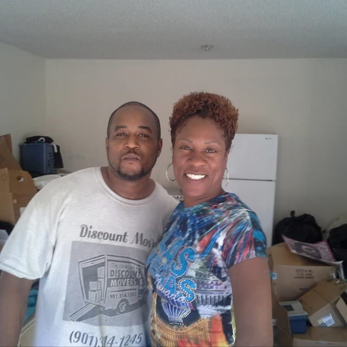 A smiling woman and a man standing together indoors, with boxes, a refrigerator, and household items in the background.
