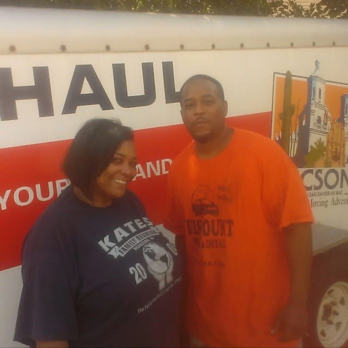 Two people standing in front of a sign that says 'HAUL'. The woman is smiling and wearing a dark T-shirt, and the man is wearing an orange T-shirt. The sign features images of landmarks and cacti.