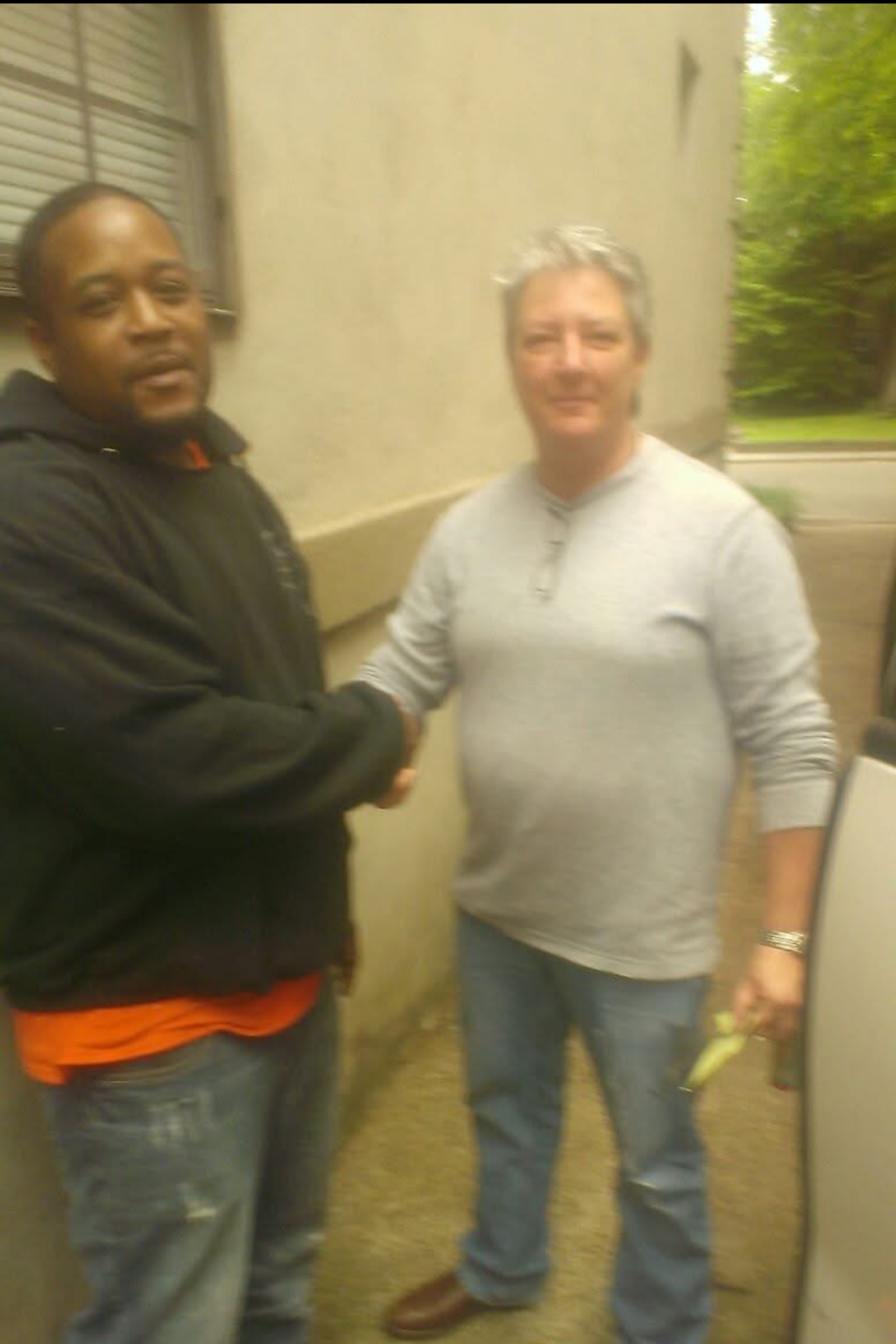 Two people shaking hands in a hallway, with one person wearing a black jacket and the other in a gray sweatshirt.