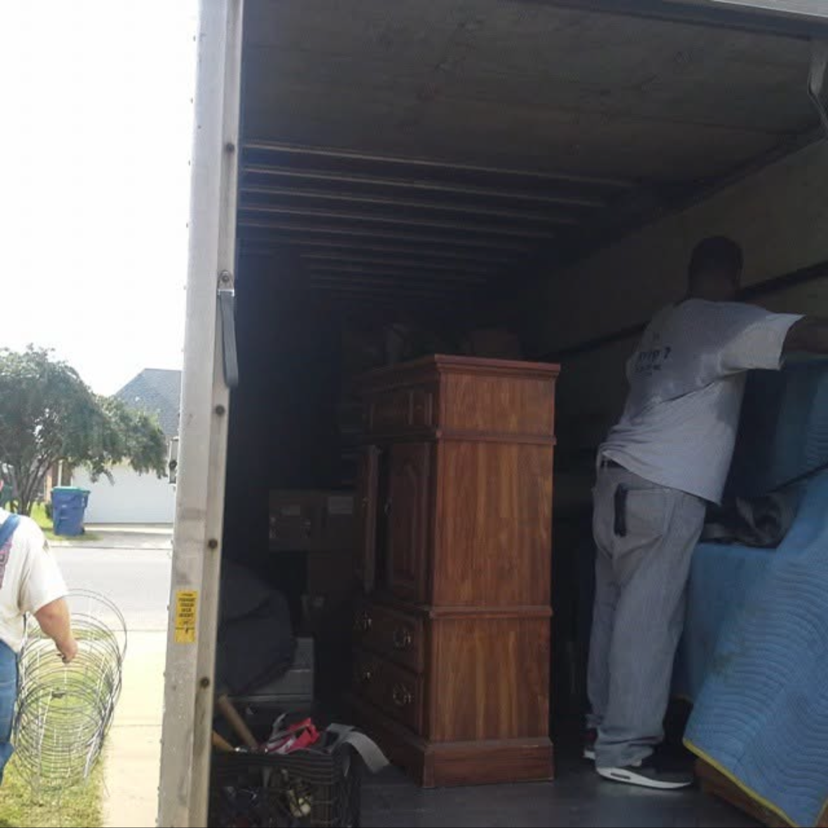 Two men packing furniture and household items into a moving truck. One man is on the right securing the load, while the other is partially visible on the left near some wire baskets.