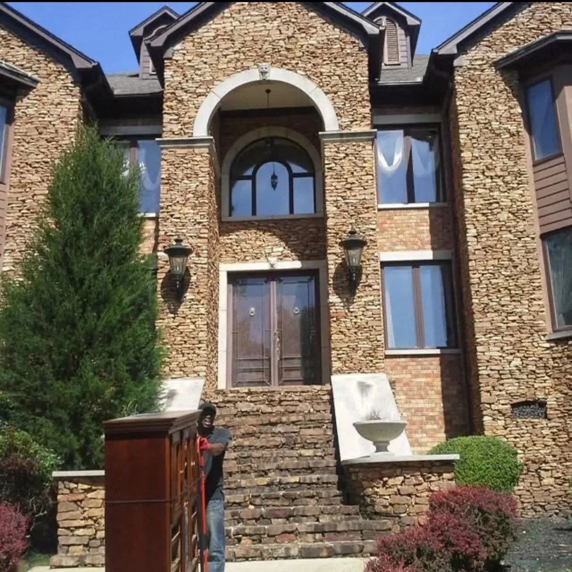 Front of a large brick house with a staircase leading to the main door. A man is standing at the bottom of the stairs near a wooden mailbox. There is greenery and shrubs in the yard, and the sky is clear.