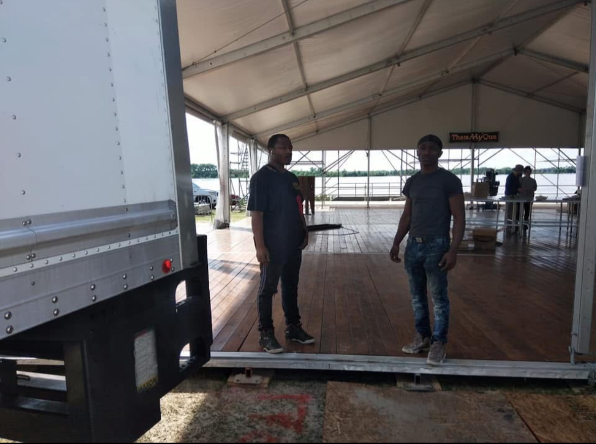 Two men standing inside a large tent structure with a wooden floor and open sides, overlooking a body of water in the background.