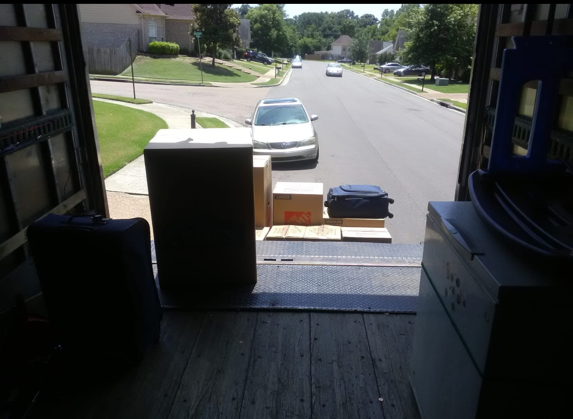 View from inside a moving truck looking out at a residential neighborhood with parked cars, green lawns, and trees.
