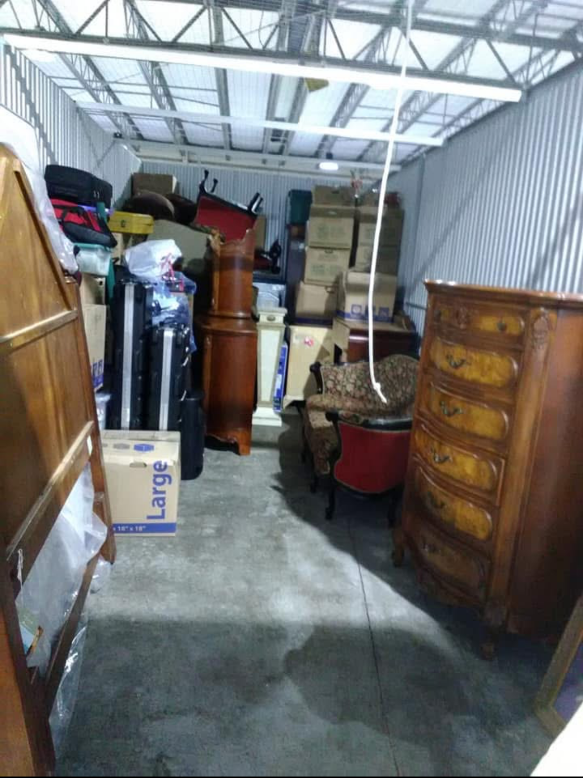 A storage unit packed with furniture and boxes, including a wooden dresser, a vintage armchair, a cardboard box labeled 'Large,' and various smaller boxes and luggage, all inside a metal-walled space with a concrete floor.