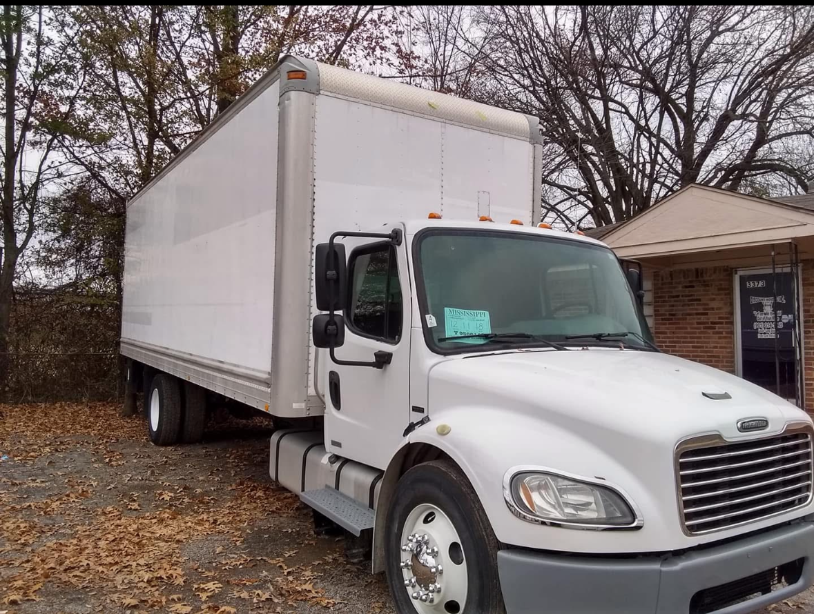 White moving truck parked on a gravel surface covered with fallen leaves, near a brick house with a porch and large trees with bare branches in the background.