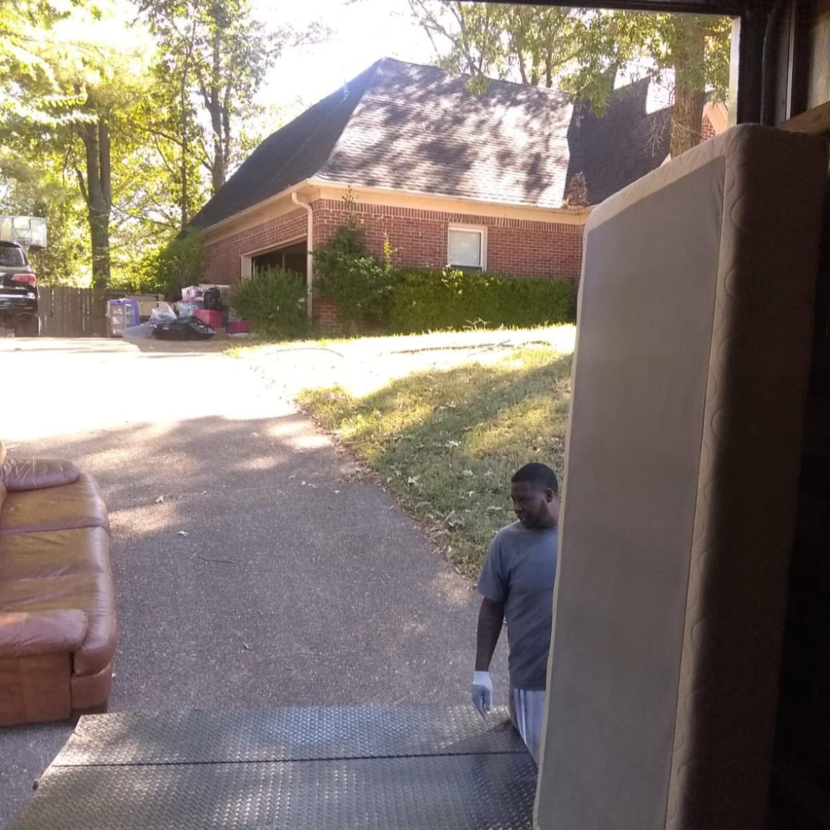 Man standing outside near a large beige object, possibly a mattress or appliance, with a driveway, a brick house, and a parked car in the background.