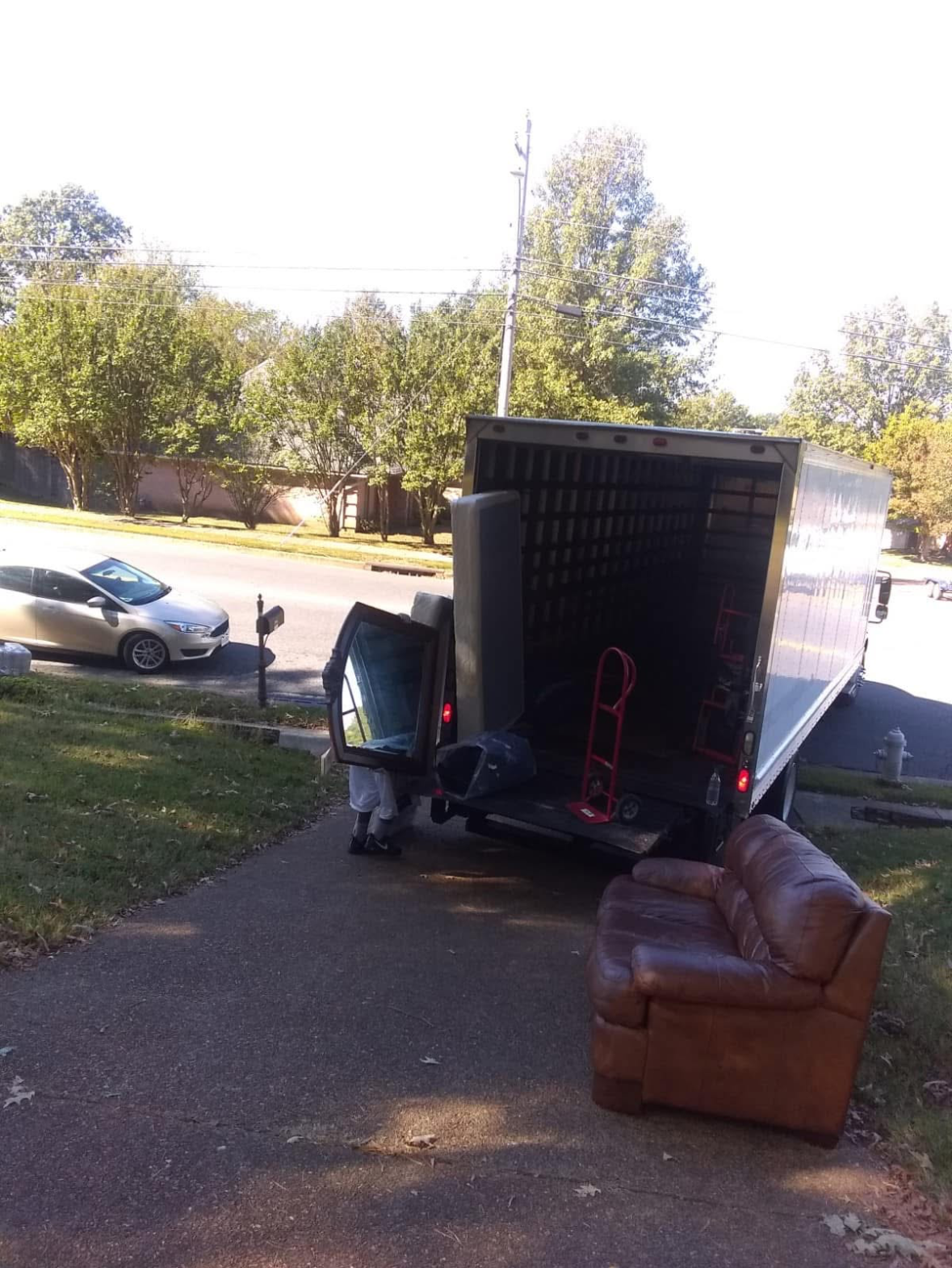 A brown leather couch is on the sidewalk near a moving truck that has its back open. A person is loading or unloading items from the truck.